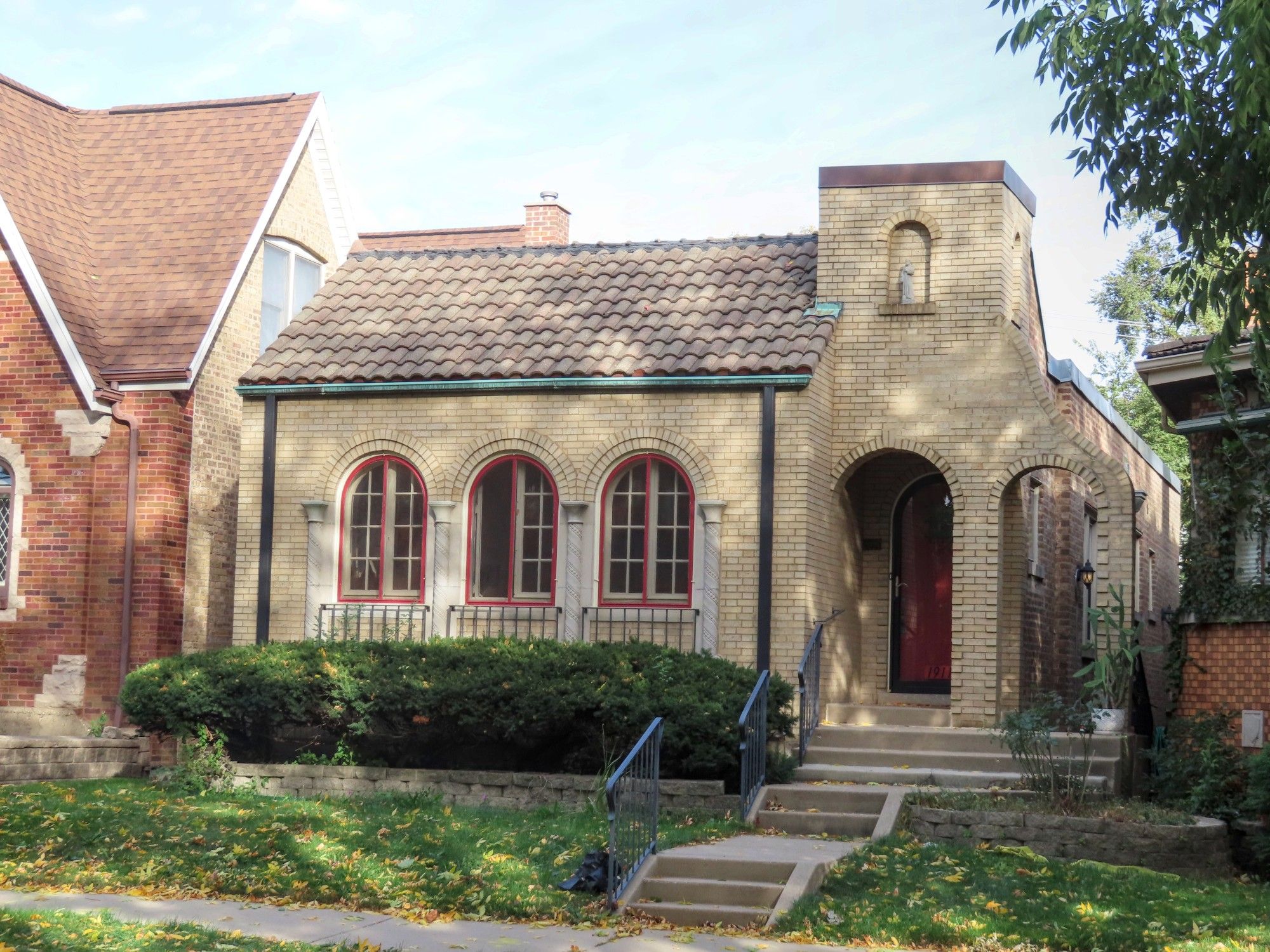 Color photo of one-story building in yellow brick. Front door is recessed behind double-arched section topped by little square tower. Most of facade is three arched windows, with stone columns in between. Roof has clay tiles.