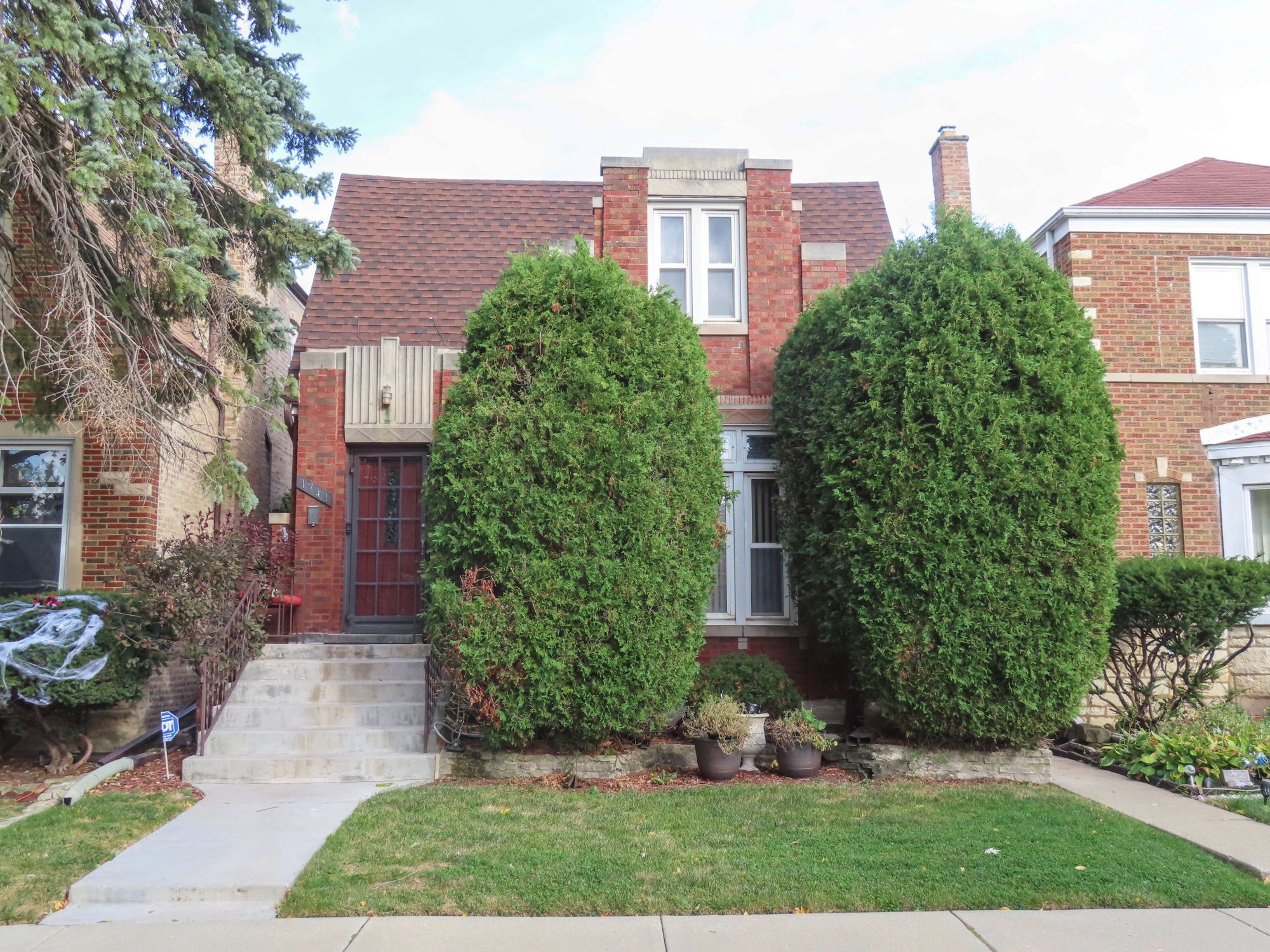 Color photo of two-story building red brick with stone trim in geometric patterns. Two overgrown bushes block sections of the building.