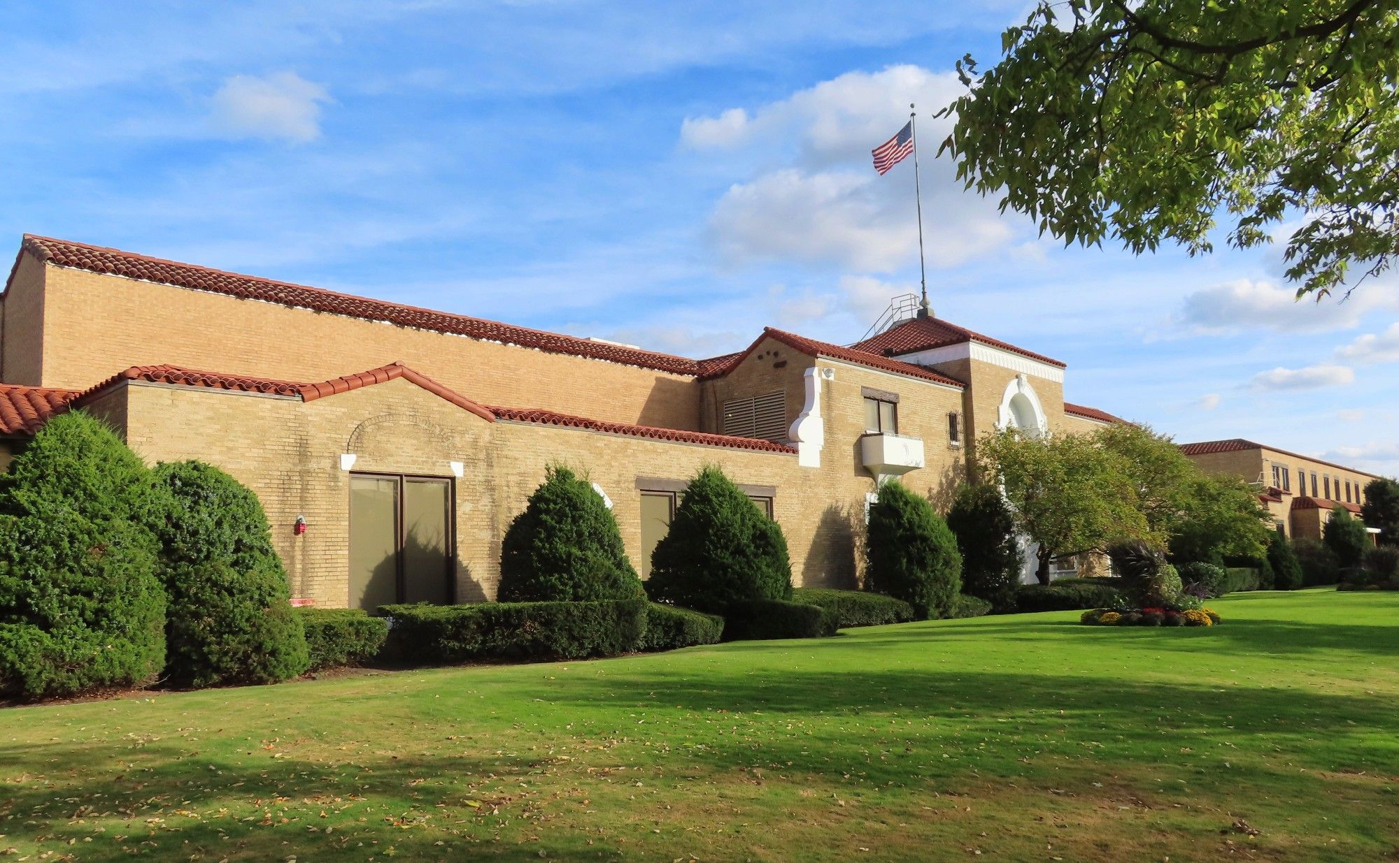 Diagonal shot of loooong mostly two-story commercial building in light brown brick, with red roof tiles. Flag flying over entrance.