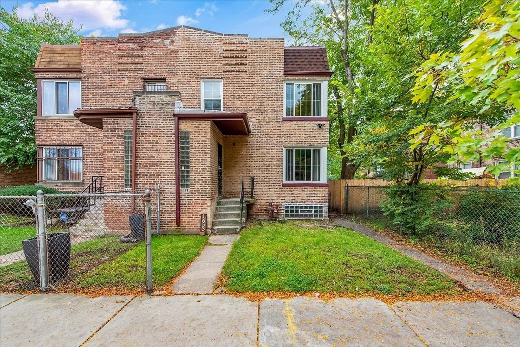 Color photo of two-story connected townhouses in common brick. Snout bay in middle for entrances with glass blocks. Subtle raised lines of brick on parapet, which rises slightly to a point.