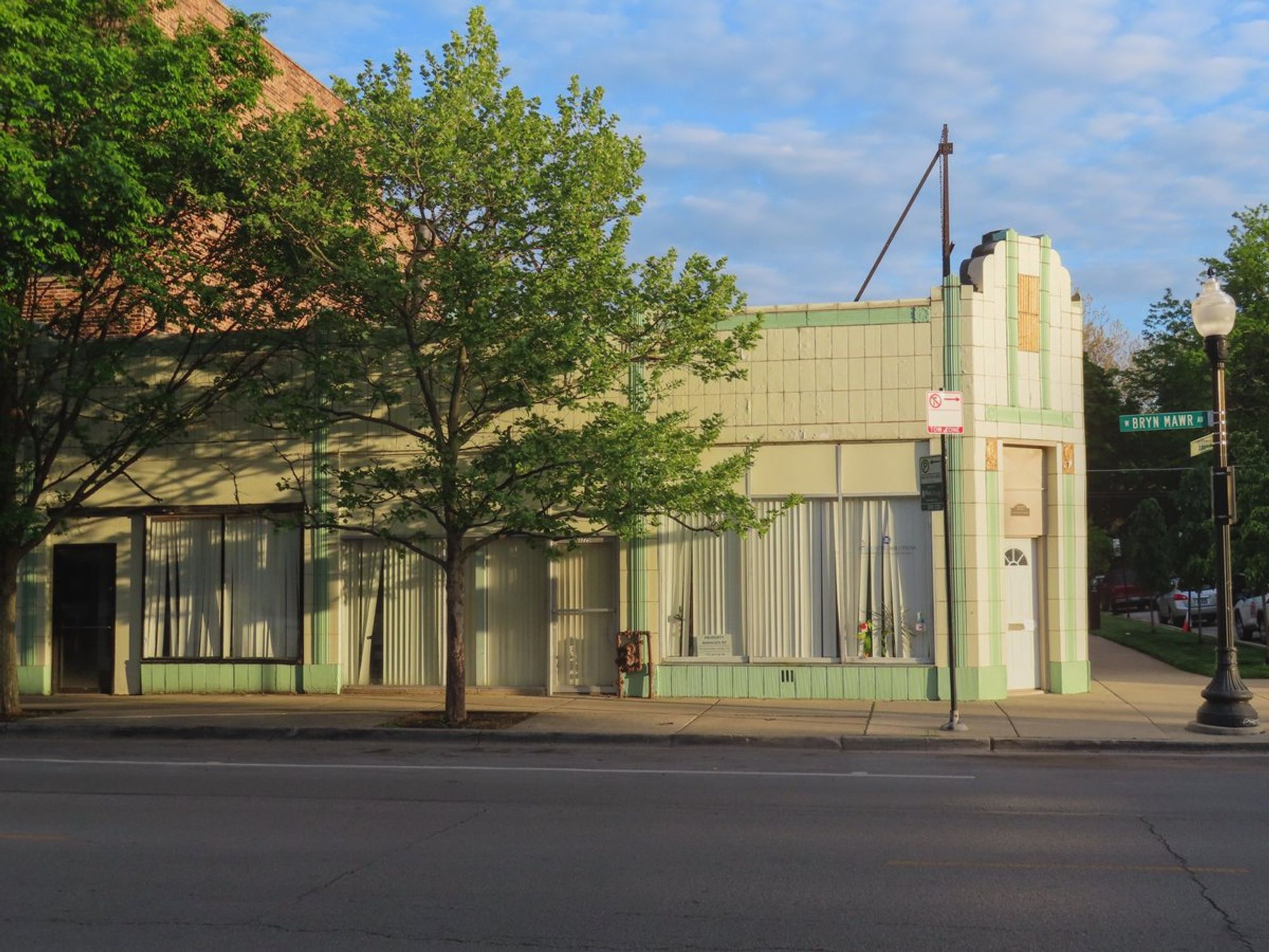 Color photo of one-story corner store clad in white terra cotta with some green trim and gold panels above the entrance.