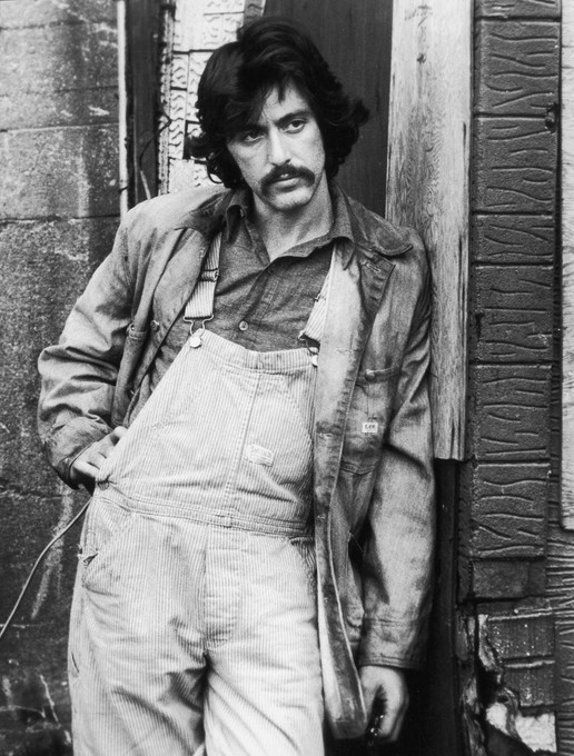 Black and white picture of Al Pacino with big hair and mustache, in work clothes, looking serious.