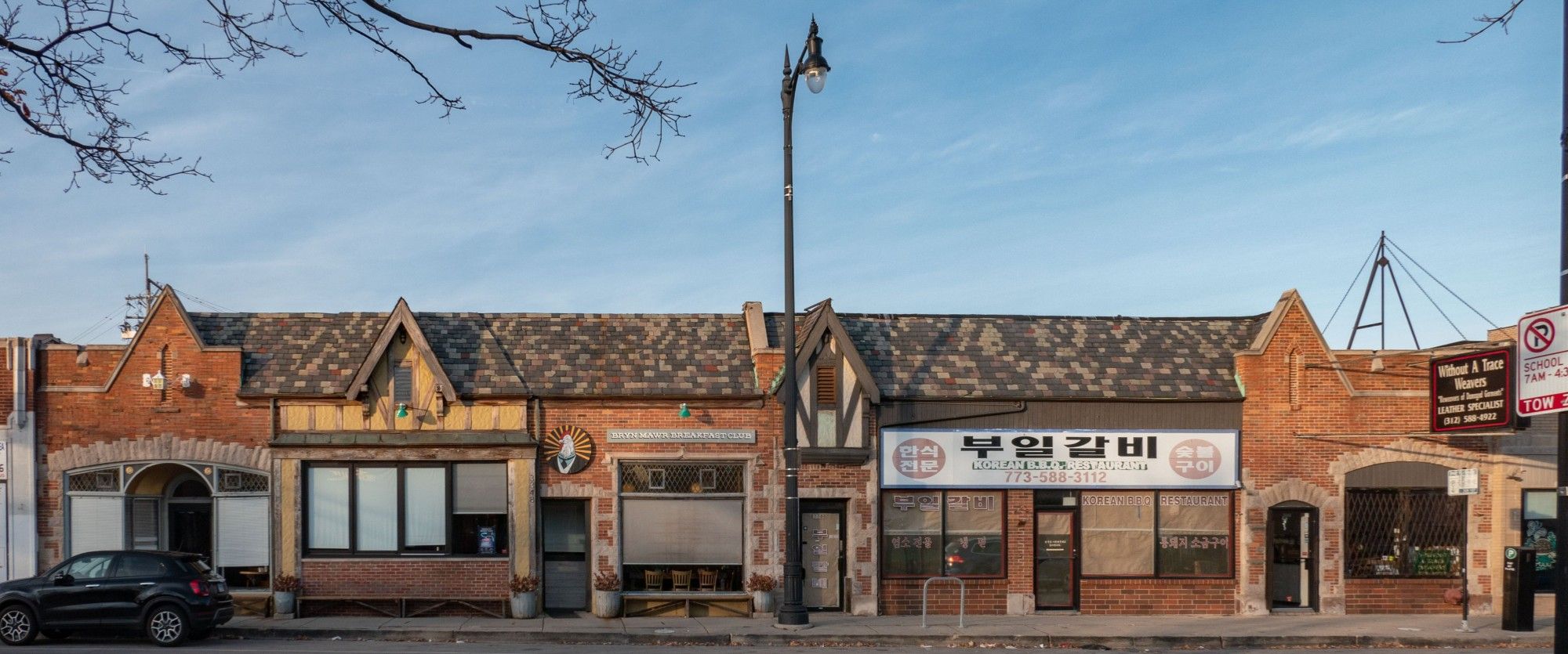 Color photo of set of connected one-story stores, connected by slate roof. Cladding includes half-timbering, red brick, and stone.