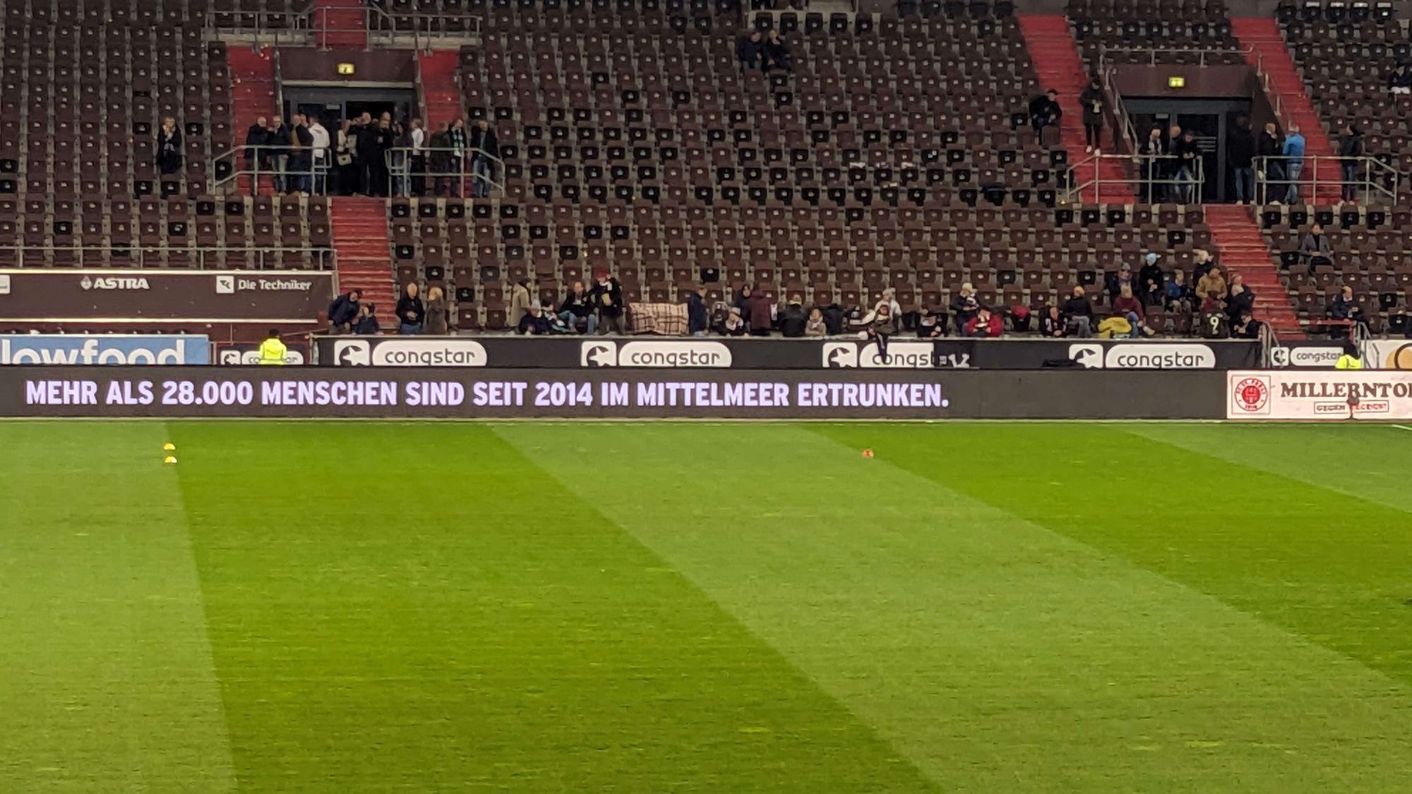 Bildbeschreibung von ChatGPT:
Das Bild zeigt eine Stadiontribüne mit leeren und teilweise besetzten Sitzen. Im Vordergrund ist ein Fußballfeld zu sehen. Am unteren Rand des Bildes befindet sich eine elektronische Werbebande, auf der der folgende Text zu lesen ist: "MEHR ALS 28.000 MENSCHEN SIND SEIT 2014 IM MITTELMEER ERTRUNKEN." Dies deutet auf eine politische oder humanitäre Botschaft hin, die auf die Tragödien im Mittelmeer aufmerksam macht. Auf der Bande sind zudem Logos von Sponsoren wie Congstar und dem Stadionnamen "Millerntor" zu erkennen, was darauf hinweist, dass das Bild im Stadion des FC St. Pauli aufgenommen wurde.