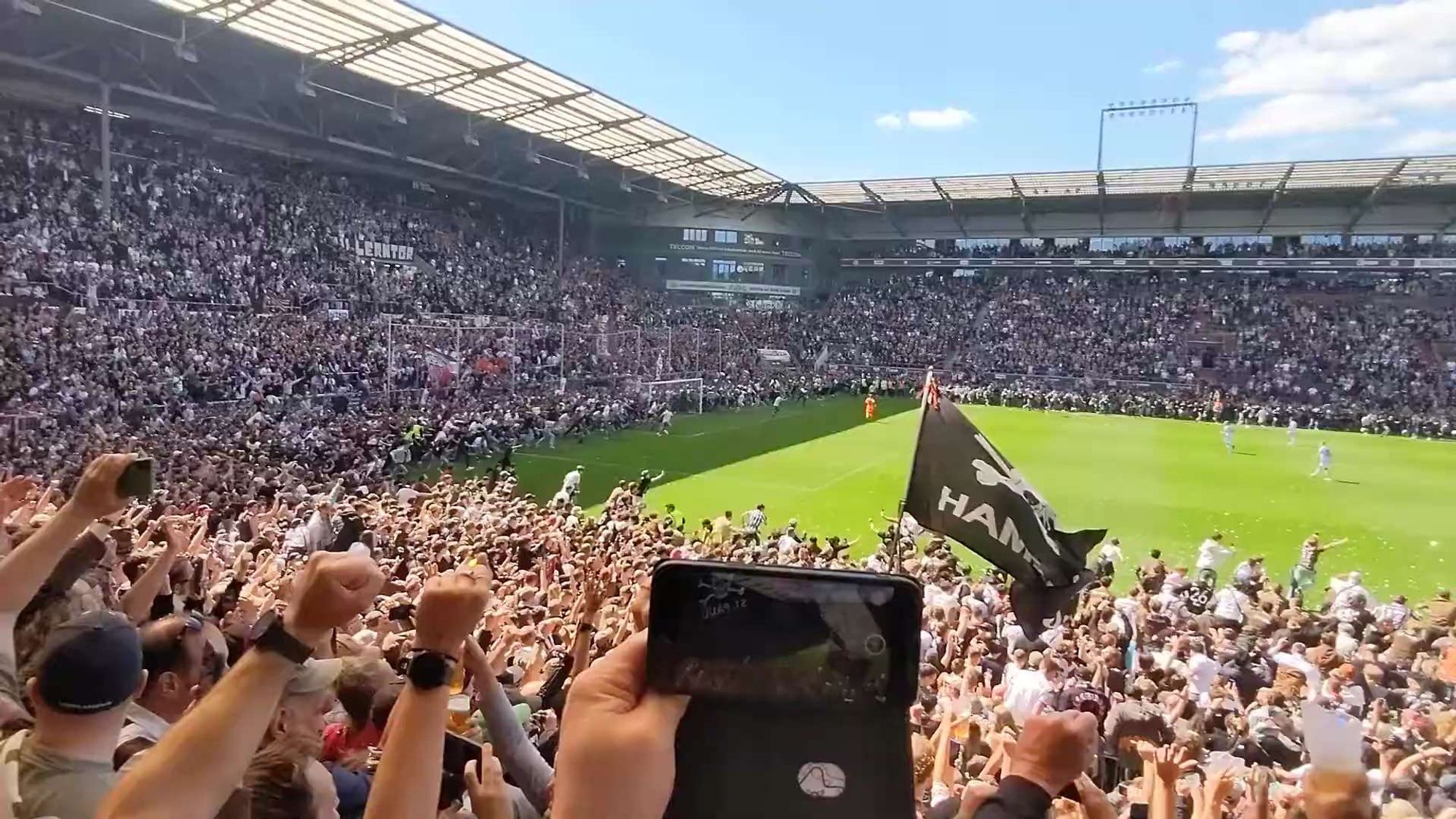 Bildbeschreibung von ChatGPT:
Das Foto zeigt eine Szene aus einem Fußballstadion während eines Spiels, wahrscheinlich im Millerntor-Stadion des FC St. Pauli. Die Tribünen sind mit tausenden von Fans gefüllt, die ihre Arme in die Luft strecken und jubeln. Einige Fans schwenken schwarz-weiße Fahnen, eine davon mit dem charakteristischen Totenkopf-Logo des Vereins. Im Vordergrund hält jemand ein Smartphone hoch und filmt das Geschehen. Auf dem Spielfeld sieht man Spieler, während auf den Rängen im Hintergrund Transparente und Fahnen hängen. Die Atmosphäre wirkt ausgelassen und voller Energie, typisch für eine leidenschaftliche Fankultur.