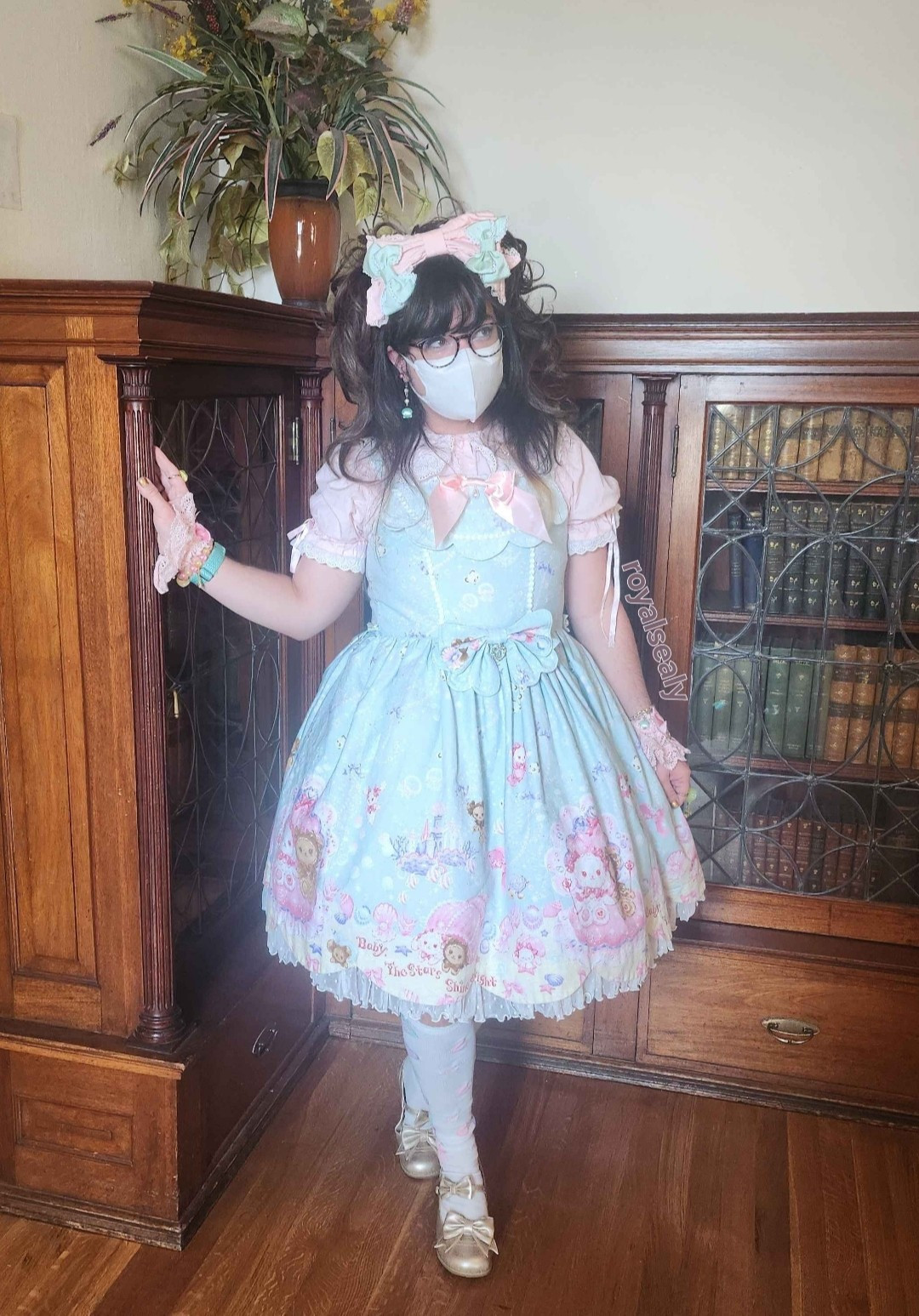 Royalsealy stands center of a book shelf, legs crossed and looking away as if in a wistfully thought. He has thick round glasses and a face mask covering the bottom half of his face.His dark brown hair is pulled up into two masses of curly hair, with a large pink headbow and mint ribbon clips in the center. On one side the hair is pulled back, allowing the viewer to see a dangly mint green shell and pearl earring. He is wearing a pink short sleeve blouse and pink wristcuffs layered underneath a marine themed mint jsk.  The JSK has various marine animals such as turtles and angelfish, and kuma kumya + usakumya as different sea creatures (sea butterfly, seahorse, and so on).  The OTKs are white with pink, mint, and lavender hearts. He is wearing gold tea parties with two bows.