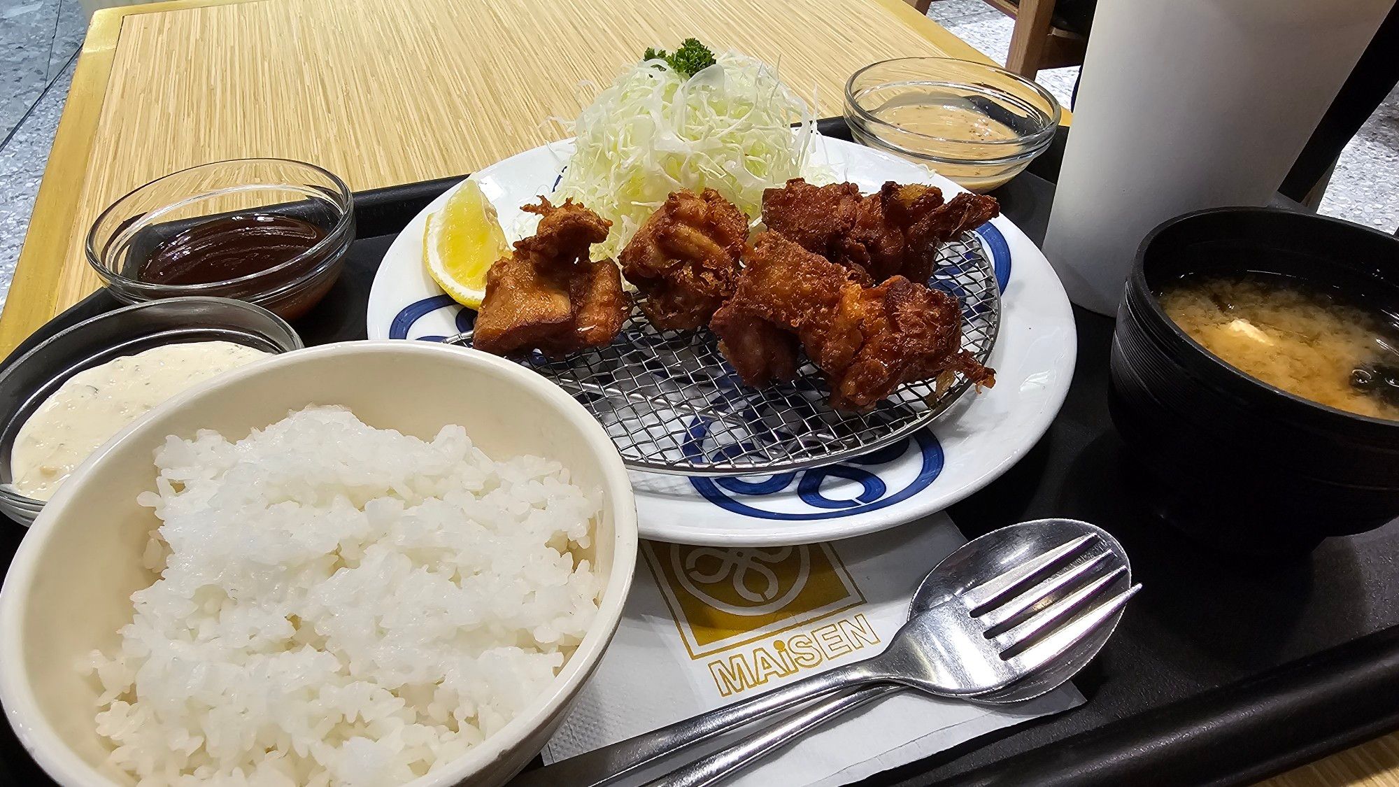 Chicken karaage on a metal mesh with a heap of finely shredded cabbage, a lemon wedge, a cup of rice, a cup of miso soup, and some dipping sauces and sesame dressing. Plus a paper tumbler of calamansi drink