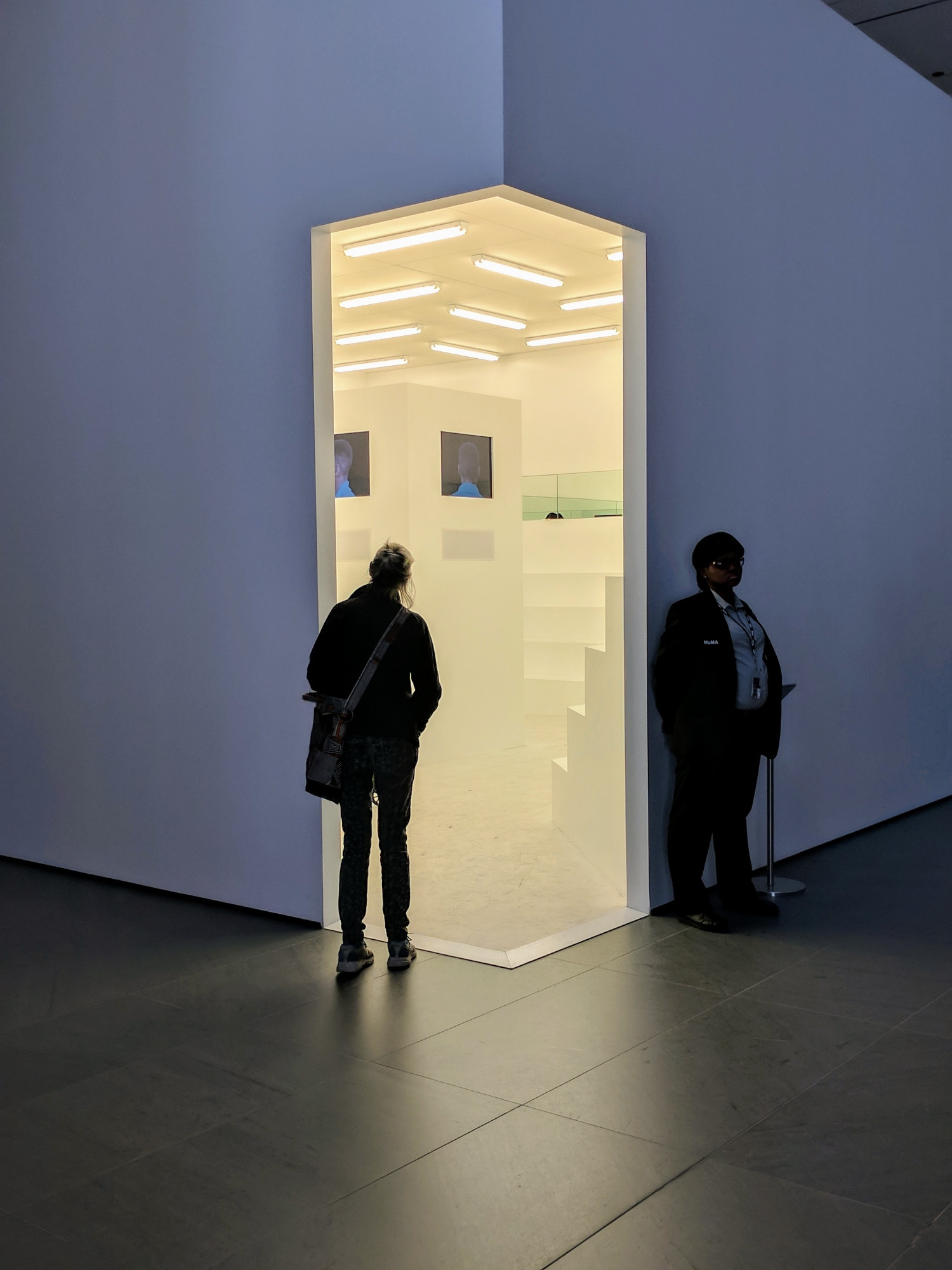 A woman looks through a door to an exhibit at the MOMA in NYC. The door and gallery inside are brightly lit while the entry is dim. A security guard stands outside the door in shadow. 
