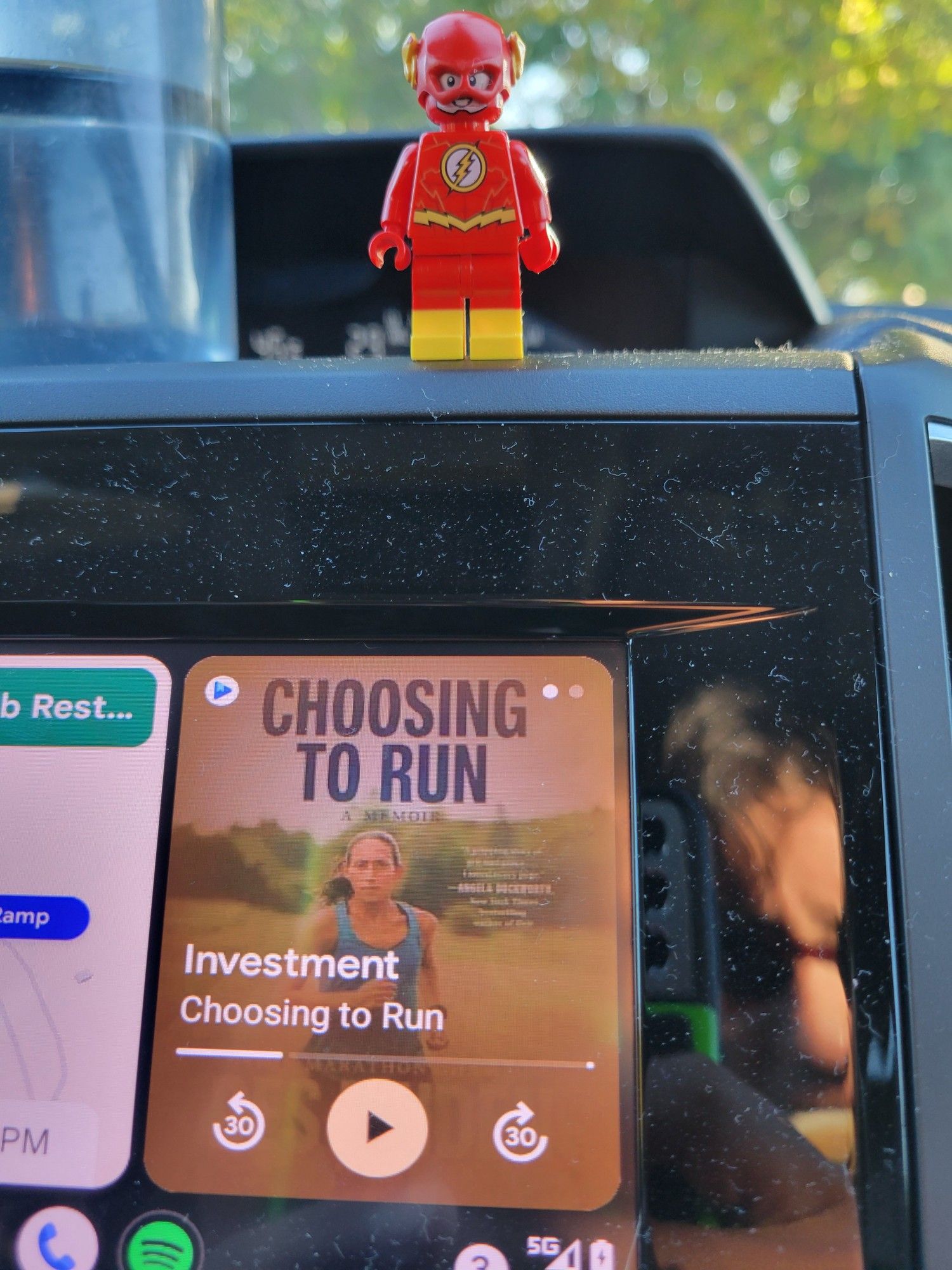 Lego Flash, a Lego mini figure of the superhero The Flash, standing on top of a car dashboard where the screen shows the audiobook we're listening to, Choosing to Run by Des Linden