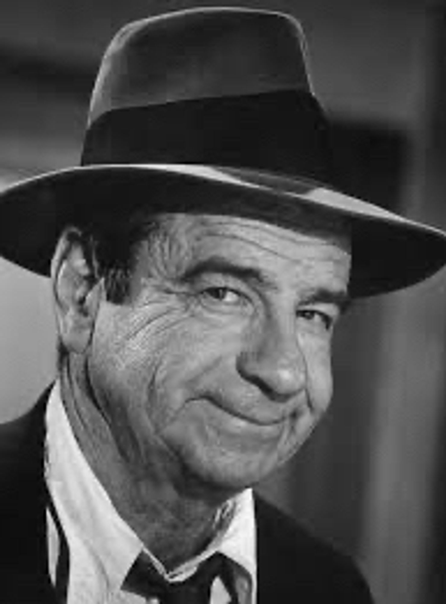 Black and white photo of actor Walter Matthau smiling at the camera. It’s a head and shoulders shot. He’s wearing a suit and old fashioned, long brimmed, fedora- style hat.