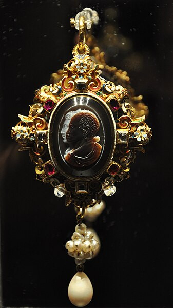 The front of the pendant features a cameo in which the brown and white sardonyx layers were cut to show a Black man and a White woman, both in profile. The enameled frame is set with rubies and diamonds, with pearl drops below.