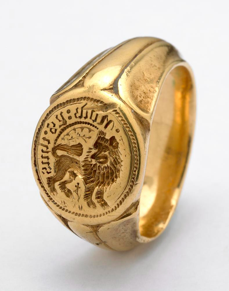 A gold signet ring with a large band, the size that you'd expect to be worn by a man. The engraved bezel features a lion standing on all four paws and gazing at the viewer.  Above the lion there's an inscription in black letters: NOW:YS:THUS, or "now is thus."