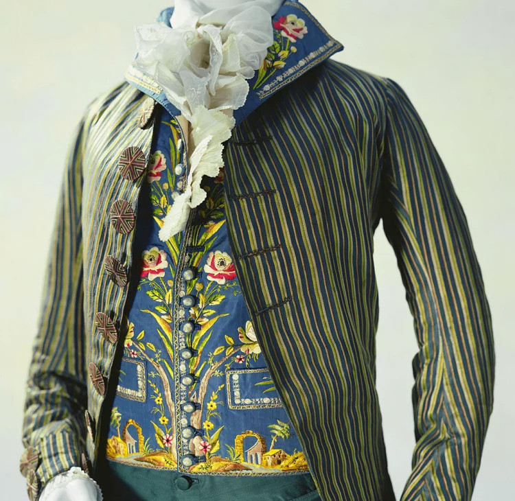 The Kyoto Costume Institute has the jacket, waistcoat, and trousers. This photo shows the jacket and waistcoat and just an inch or two of the green trousers' waistband. The jacket is satin, with blue and green vertical stripes, a fold back standing collar, and buttons decorated with angular geometric patterns. And that waistcoat!  Vivid blue, embroidered with stems and flowers running up the center and continuing on the stand-up collar, and embroidered with ruined Roman arches and outbuildings along the bottom edge. Under the waistcoat at the neck and cuffs we see lovely white frills from the shirt underneath.