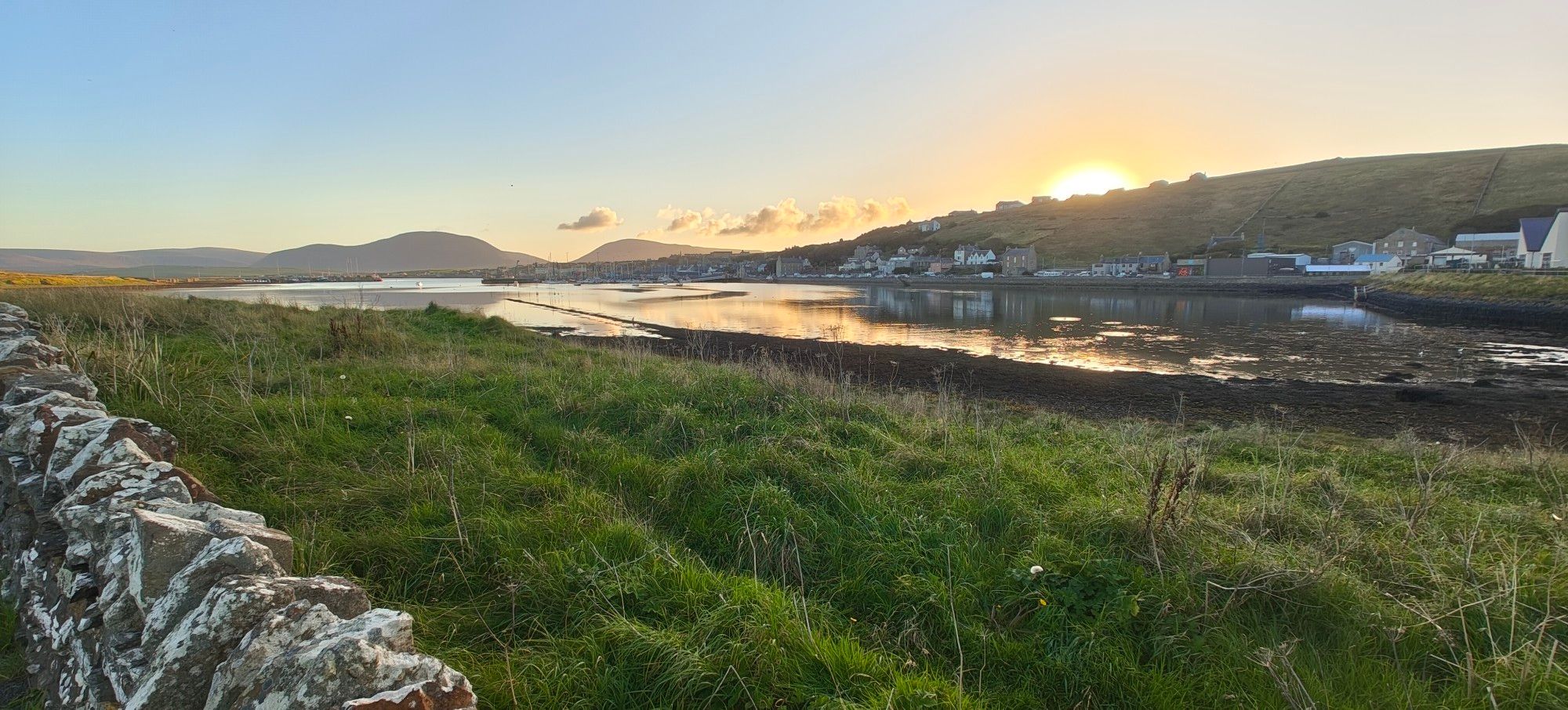 Sunset over a bay with a town on the far side. Hills in distance, grass in foreground.