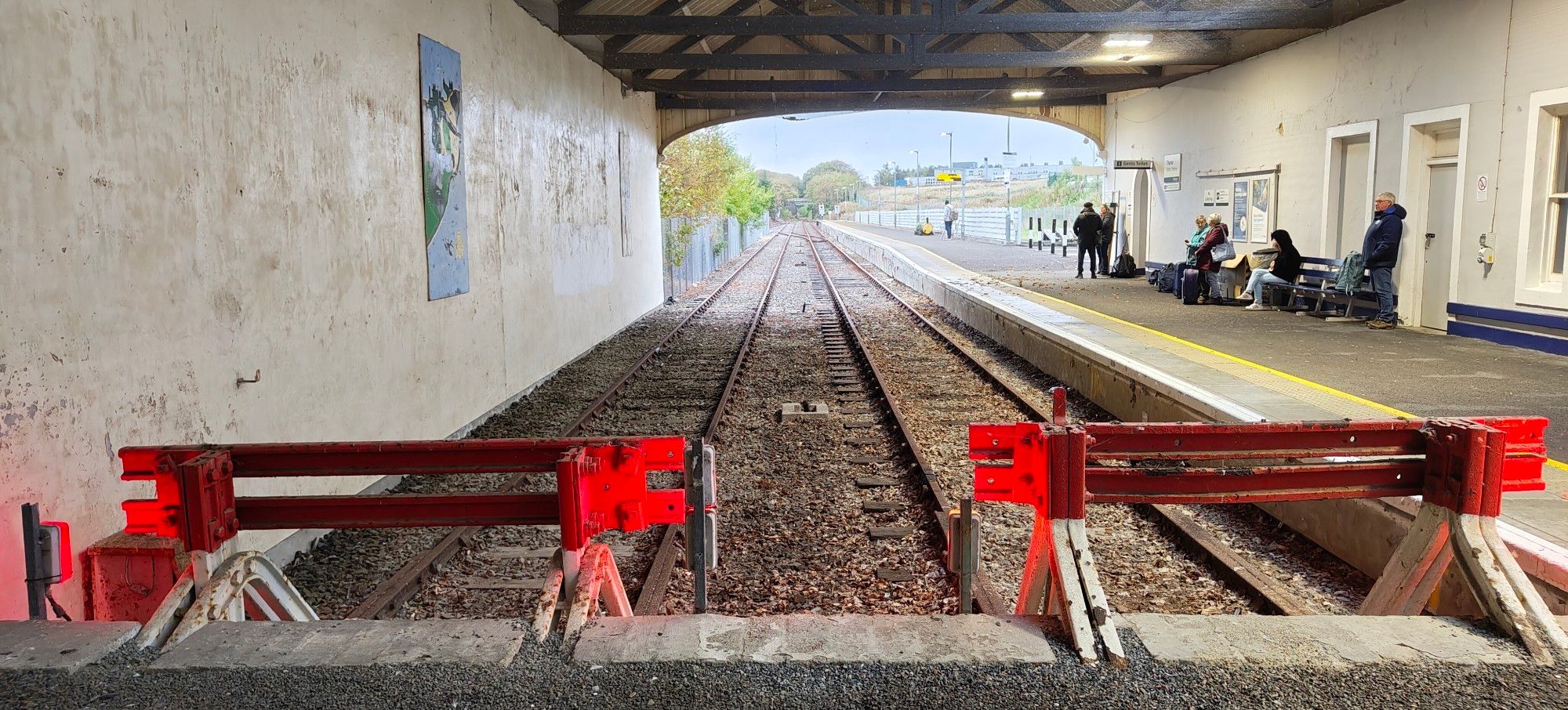 Red buffers in foreground, two rusty railway tracks beyond. A single platform, and  a basic roof overhead.