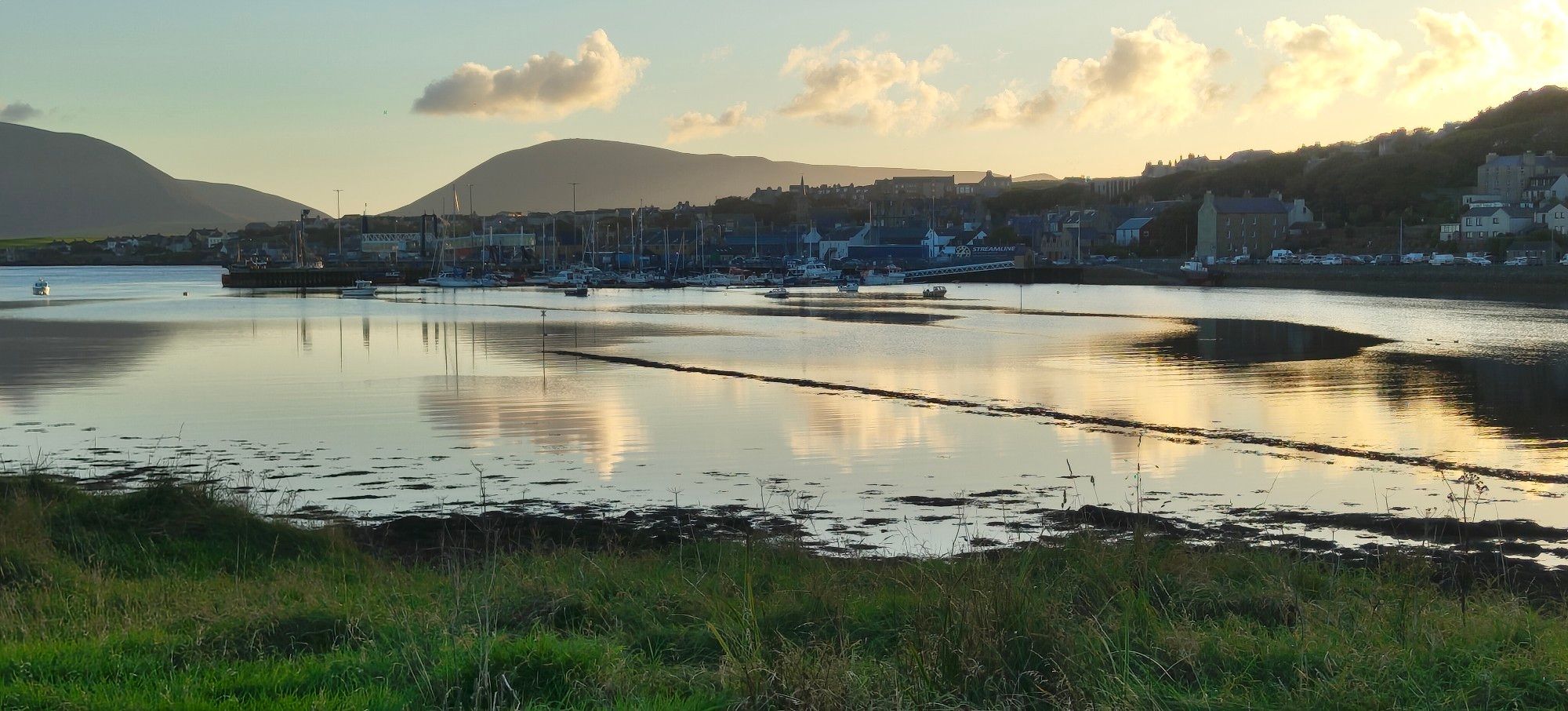 Sunset over a bay with a town on the far side. Hills in distance, grass in foreground.