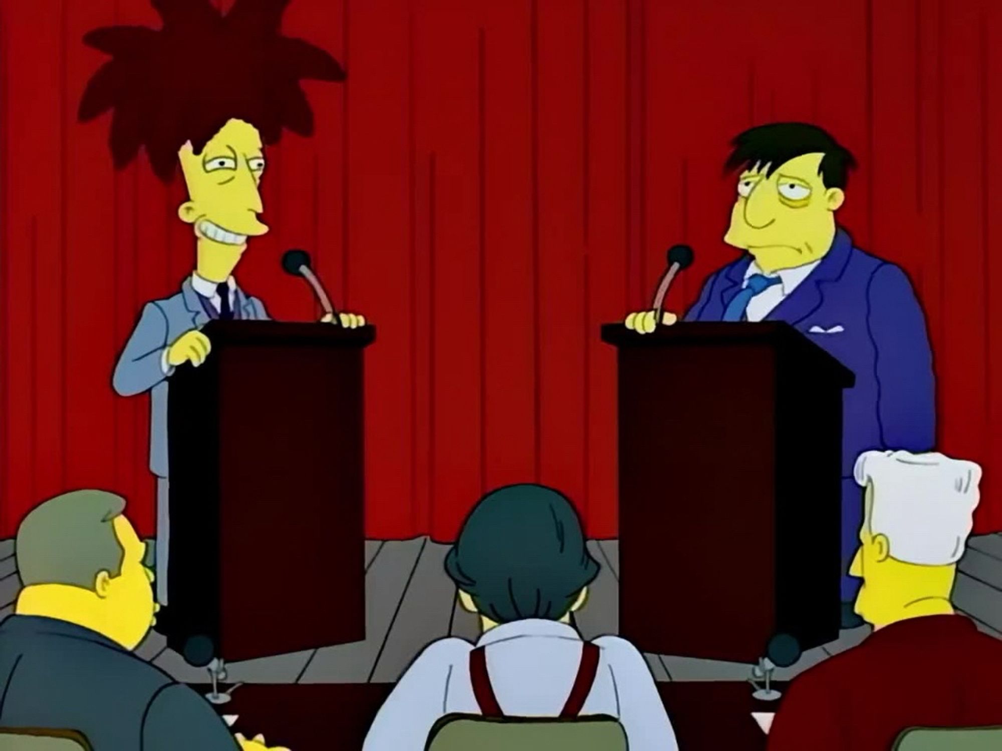 “Sideshow” Bob Terwilliger and Mayor “Diamond” Joe Quimby stand at lecterns on a debate stage. In the foreground we see three moderators from behind: Springfield anchorman Kent Brockman, a Larry King lookalike, and arch-conservative Springfield talk radio personality Birchibald T. “Birch”Barlow