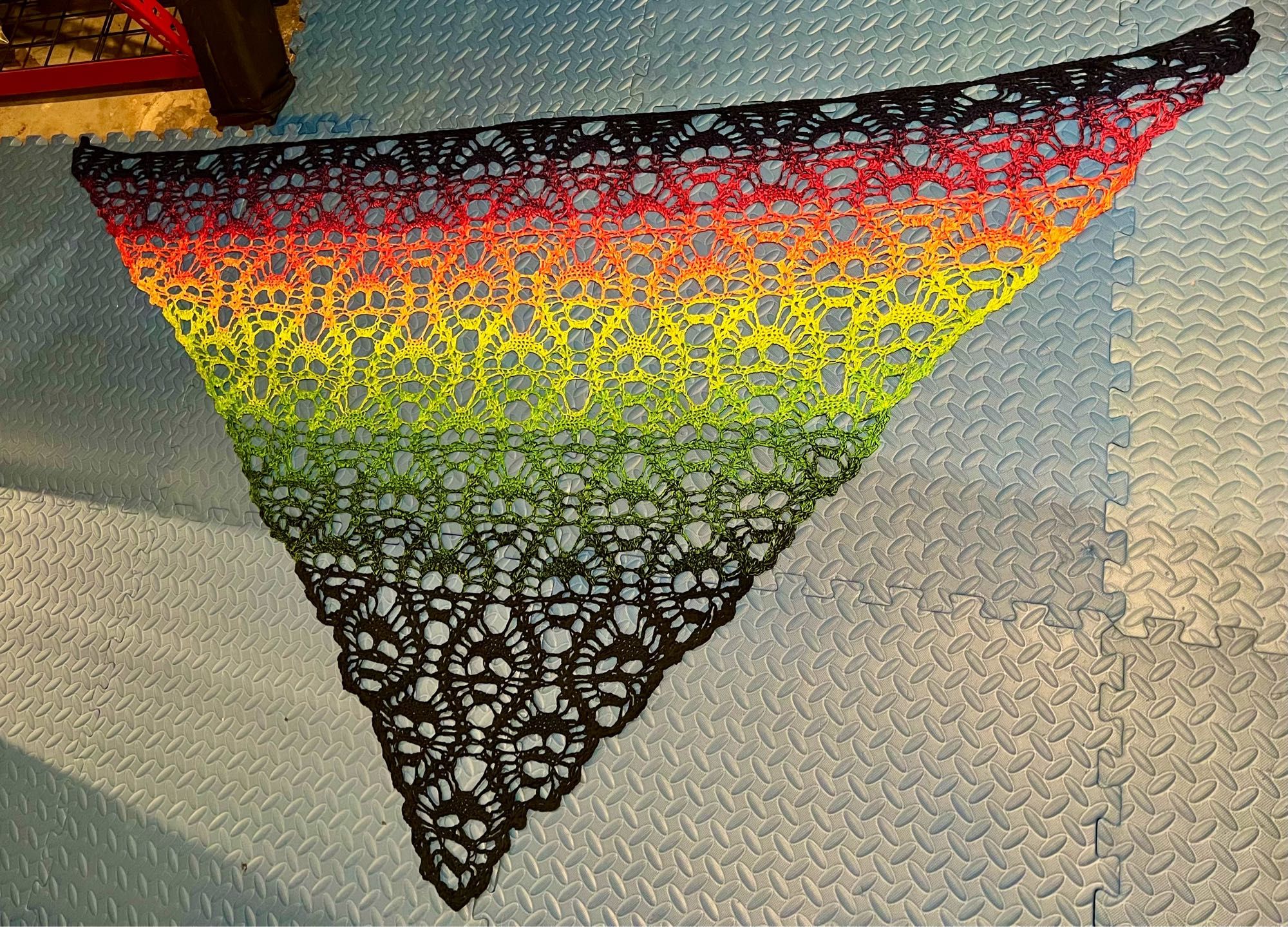 A crochet shawl spread out on blue mats, in a gradient fading from dark purple through pink, orange, yellow, green, to black. The motif is tessellated skulls.