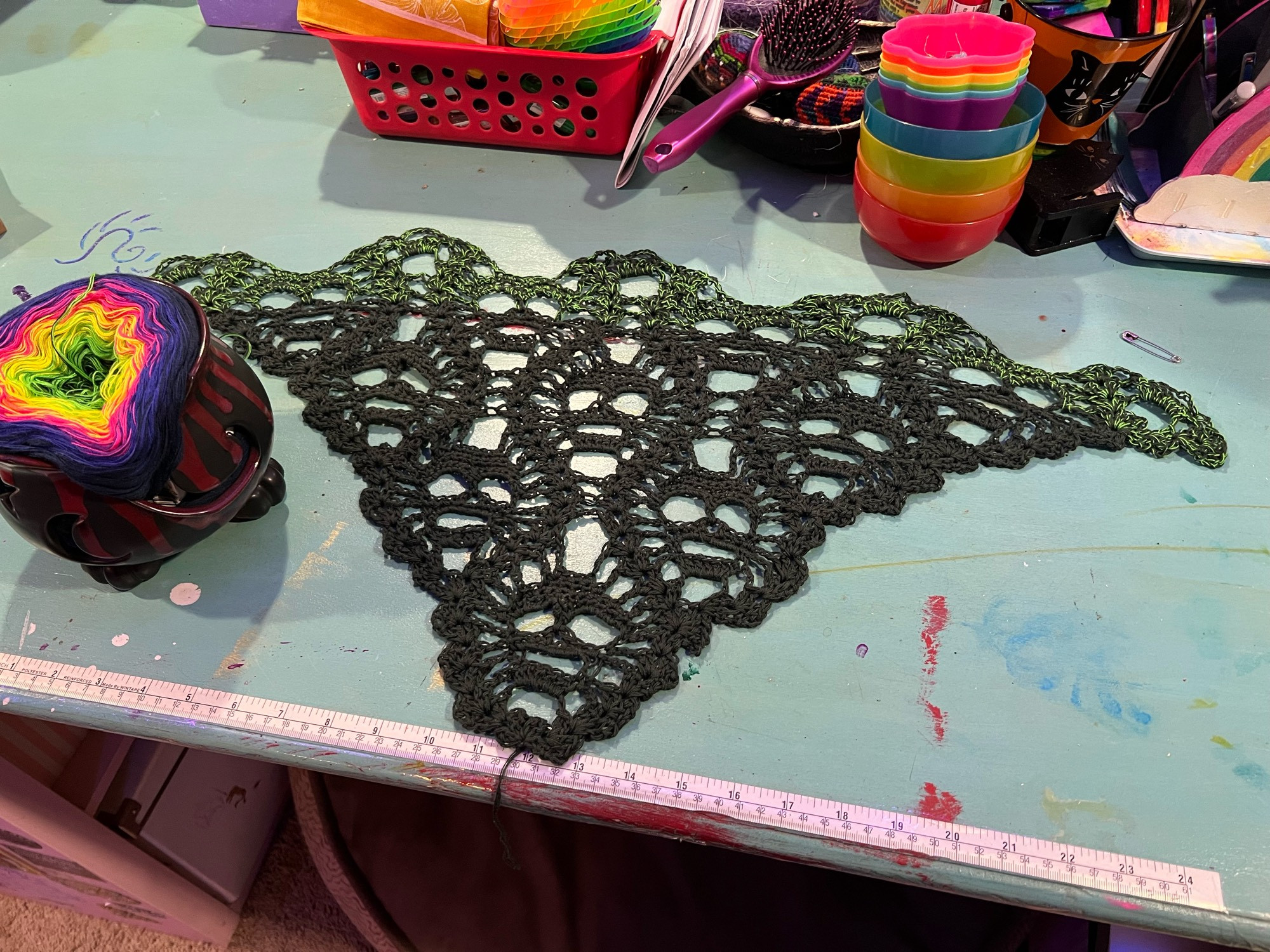 A black and dark green triangle shaped crochet piece: there is a repeating skull motif. It is on a seafoam green table, with a ceramic cauldron full of yarn.