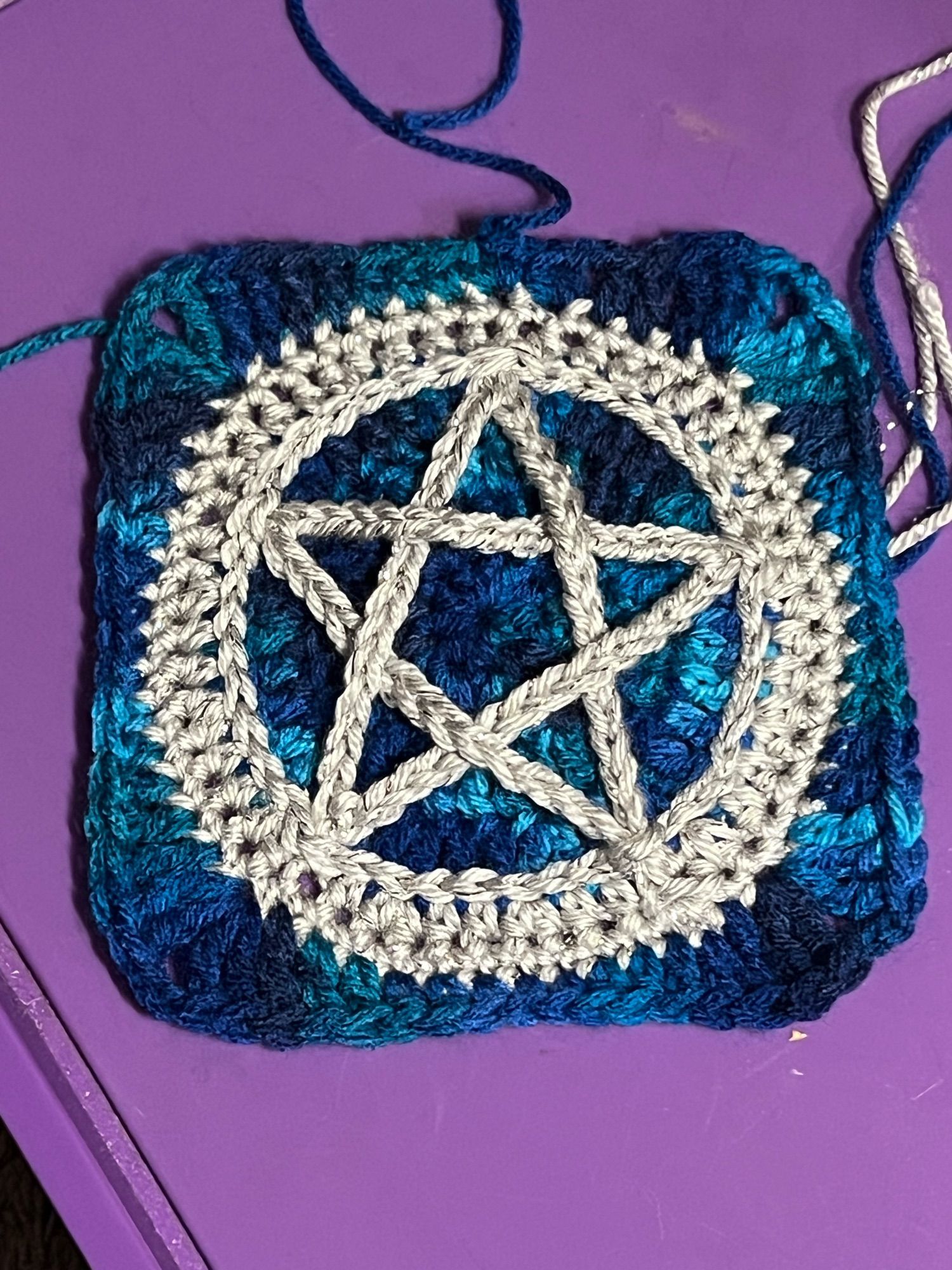 A crocheted granny square in shades of turquoises and blues, there is a silver pentacle as the center design.