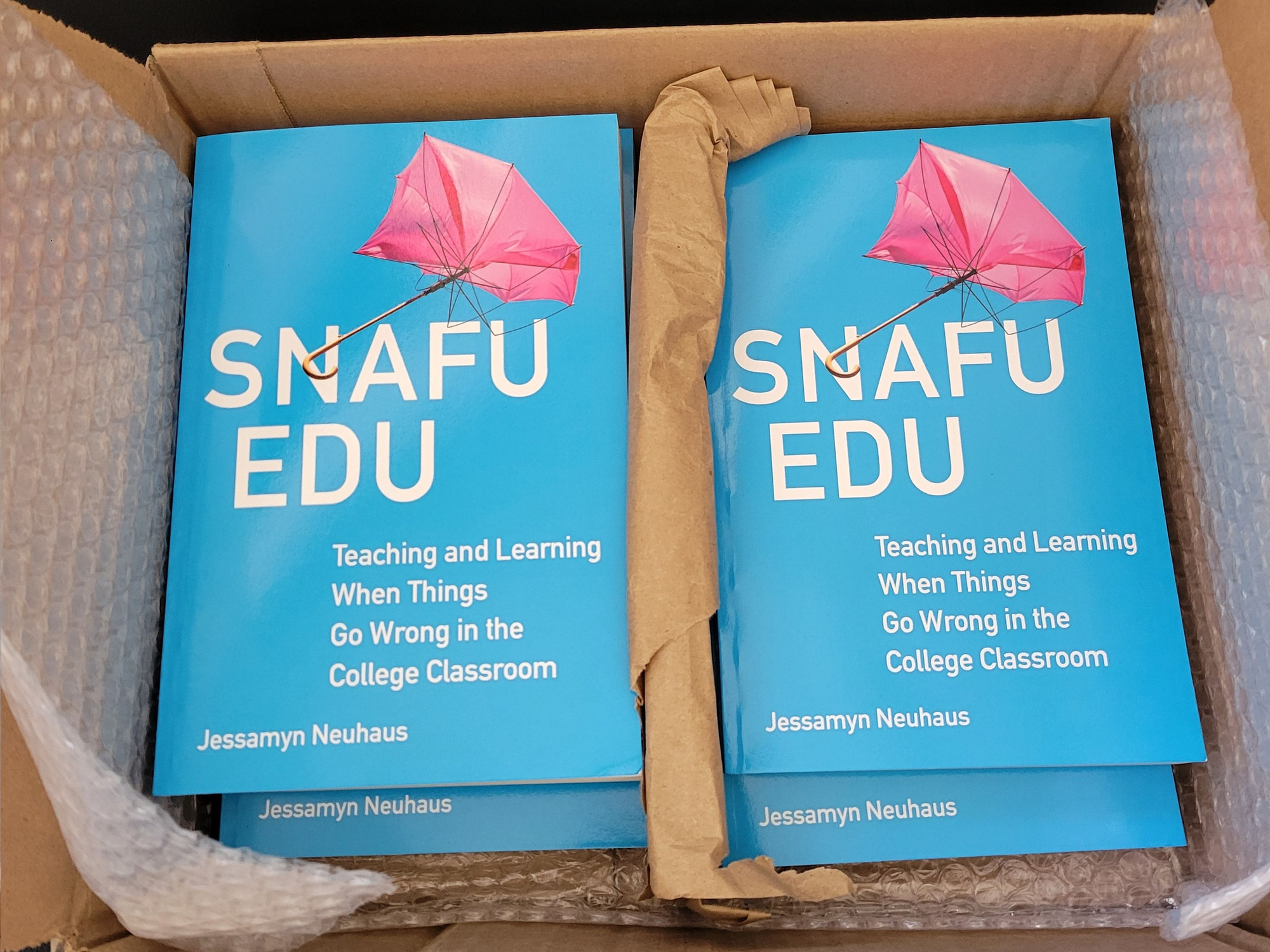 Box of books, copies of Snafu Edu: Teaching and Learning When Things Go Wrong in the College Classroom by Jessamyn Neuhaus. Cover is bright blue with an image of an inside out pink umbrella.