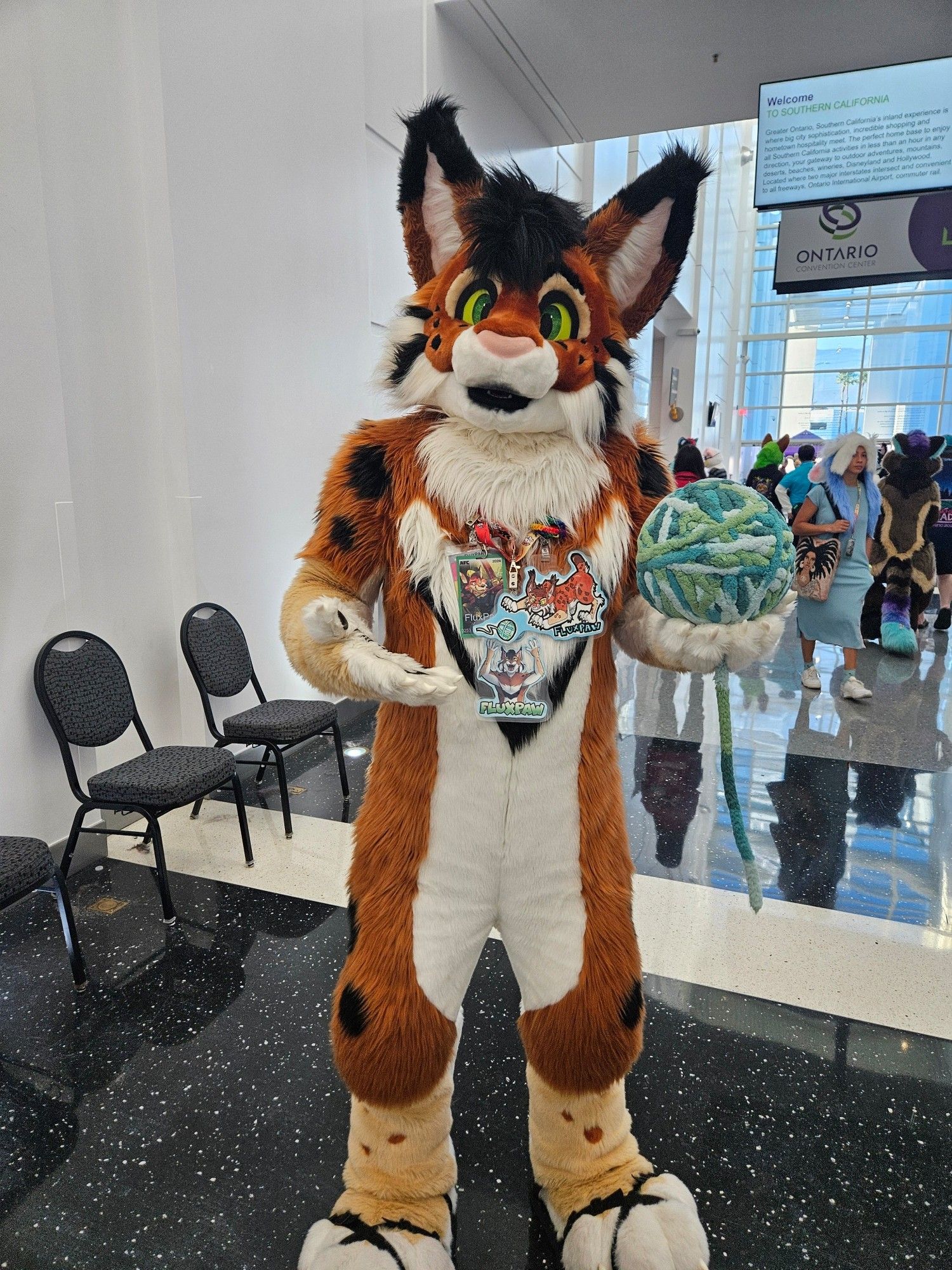 A lynx fursuit by the name of Fluxpaw. Holds a ball of yarn in one paw.