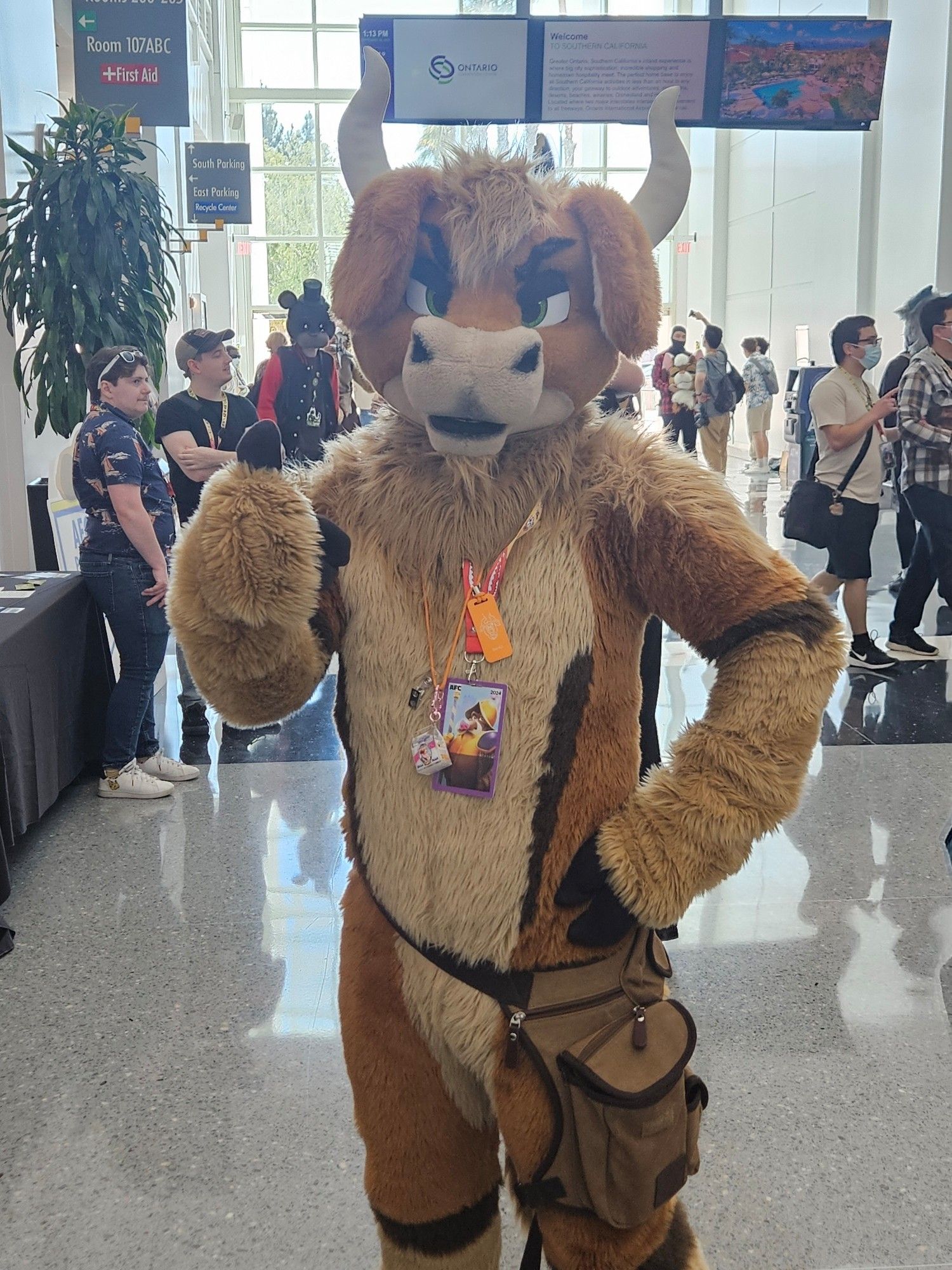 Bento the bovine fursuiter, giving the camera a thumbs up. Has badges and a carry on bag.