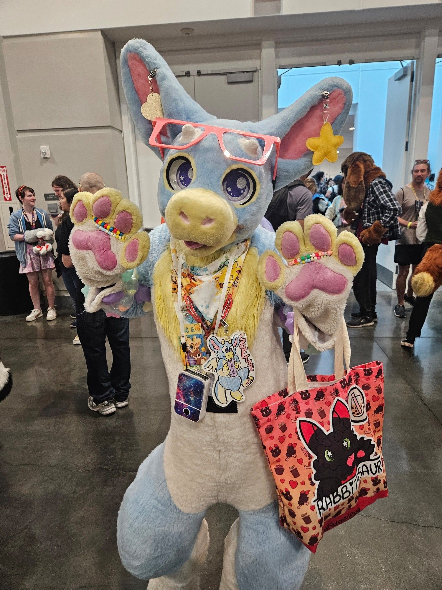 Softie the baby blue and white angle dragon fursuit with pink highlights. Both paws are up waving at the camera.