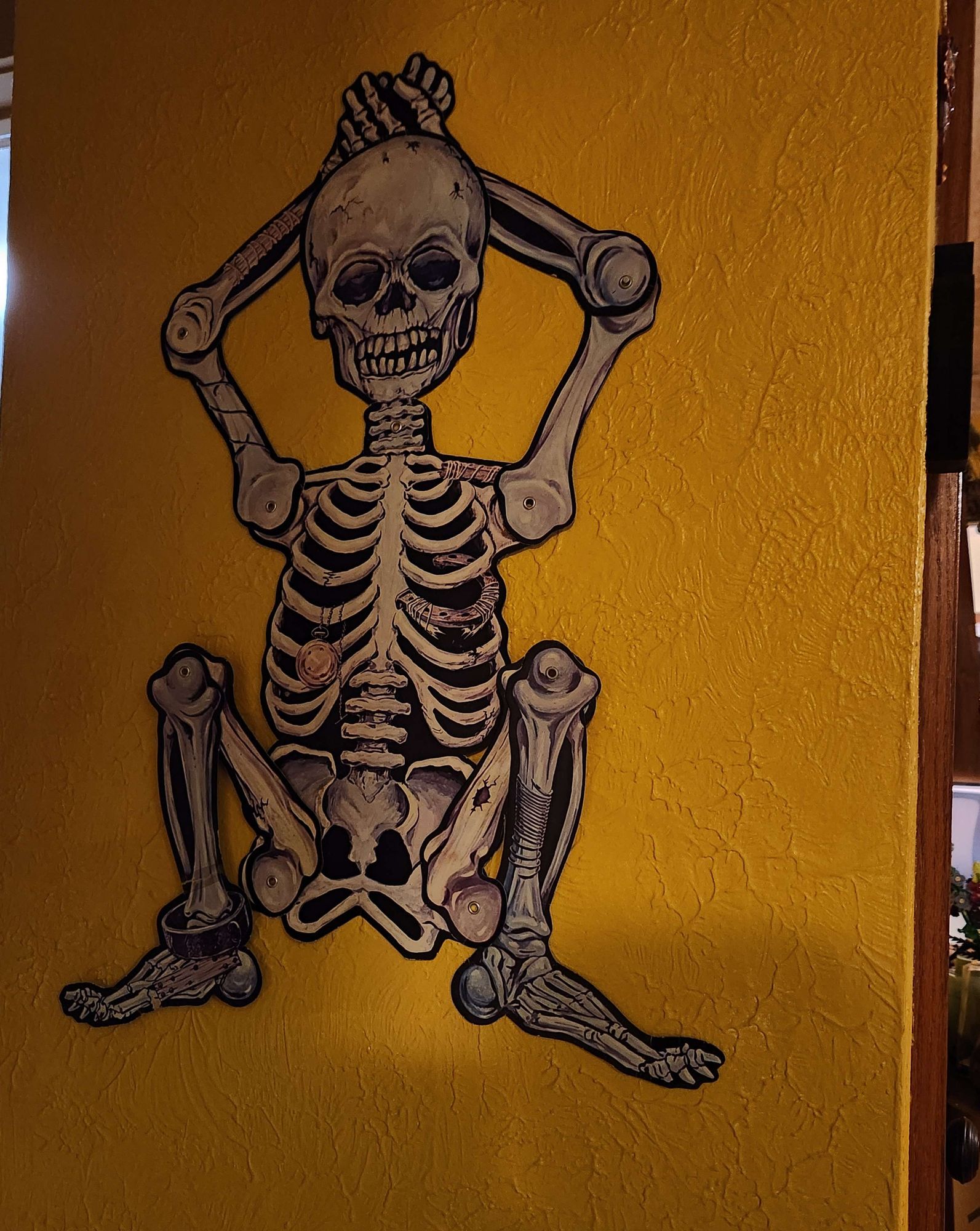 A vintage paper skeleton decoration stuck to my yellow wall. His arms are bent behind his head and his legs are folded up to strike a very scandalous pose