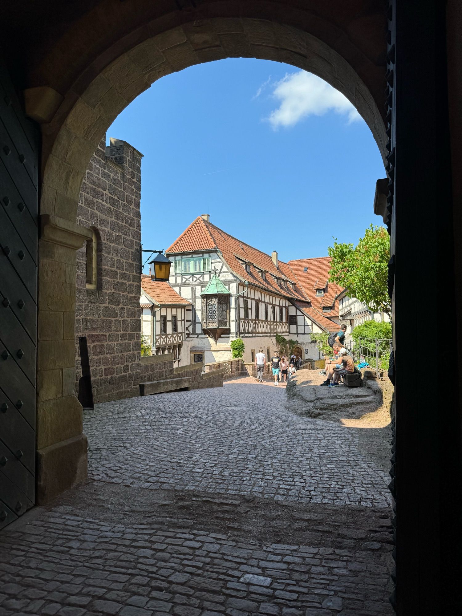 Blick durch die Torhalle mit Torbogen auf die Vorburg. Der Himmel ist beinahe wolkenlos. In der Mitte sind Fachwerkhäuser zu sehen. Der Boden besteht aus groben Pflaster. Einige Besuchende spazieren oder sitzen am Rand.