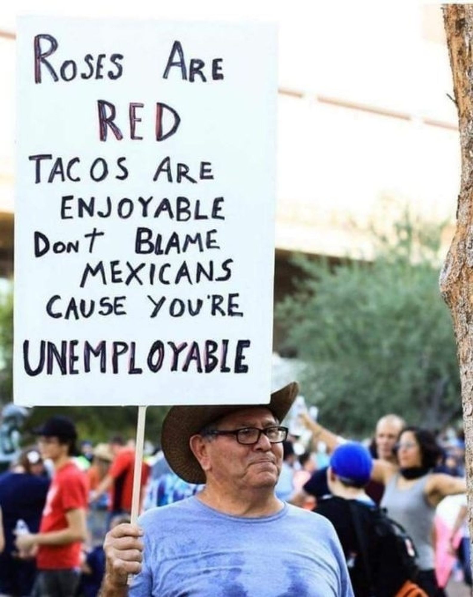 Man holding a sign that says, "Roses are red, tacos are enjoyable, don't blame Mexicans because you're unemployable."