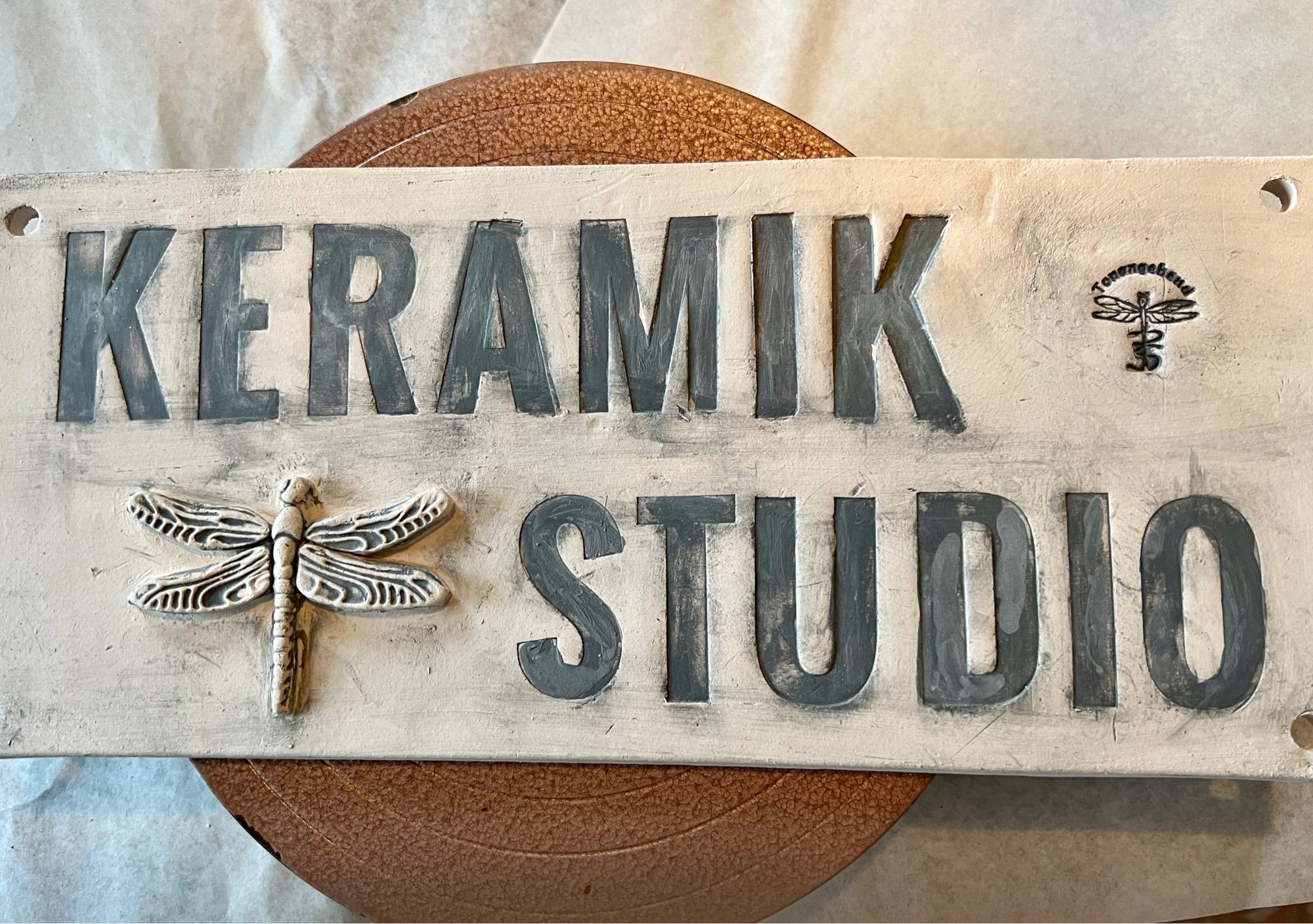 Firmenschild noch unglasiert, mit grauer Schrift: KERAMIK STUDIO Am linken unteren Rand ist eine Libelle, oben rechts der Firmenstempel Tonangebend mit Libelle und meinen Initialen PS