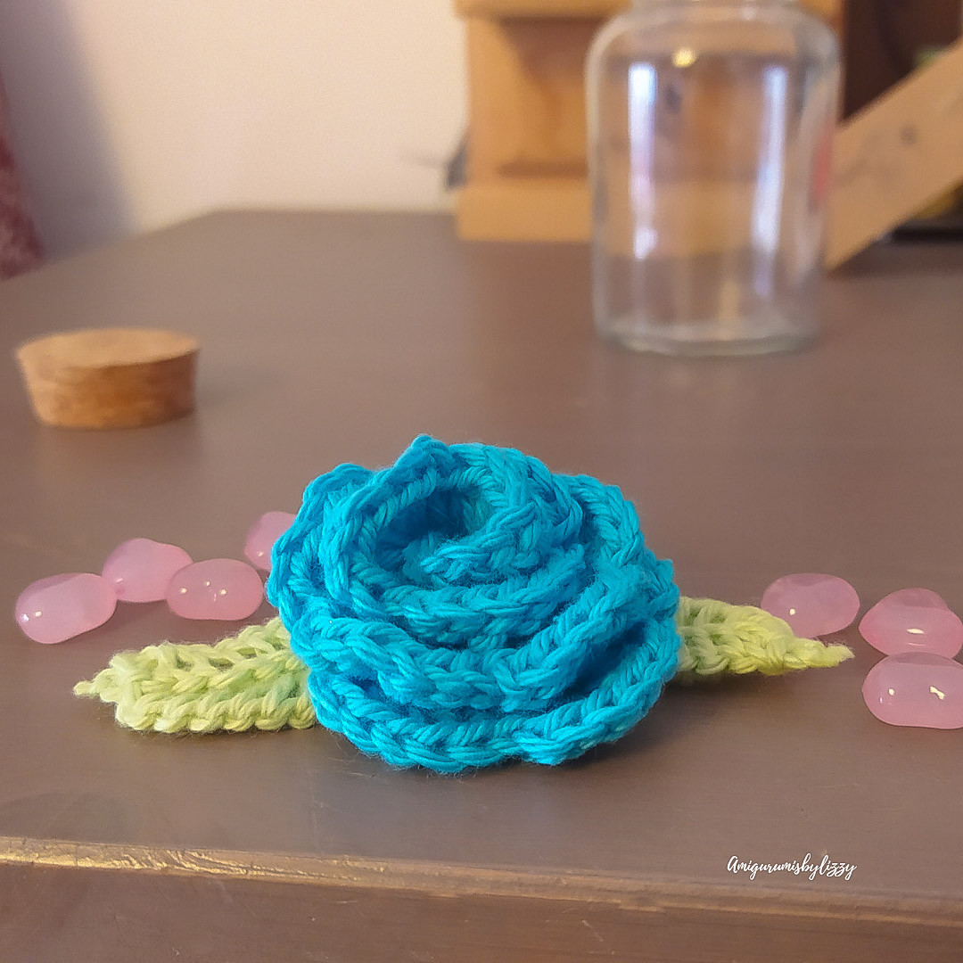 Una rosa de color azul de crochet con hojas verdes y con cuentas rosas en forma de corazón alrededor, 