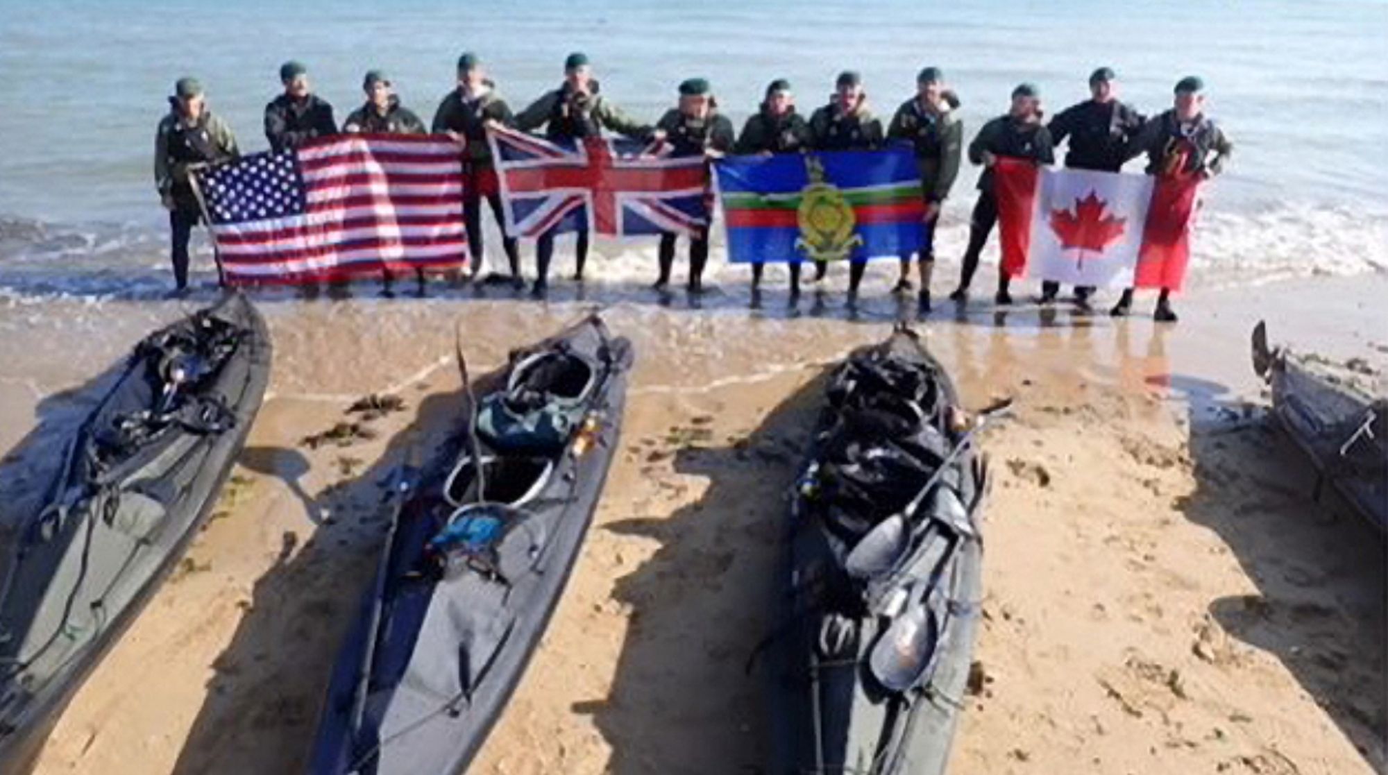 Standing as a group behind their kayaks on one of the Normandy beaches. They’re holding flags of the USA, the UK and Canada, and the Royal Marines’ flag.