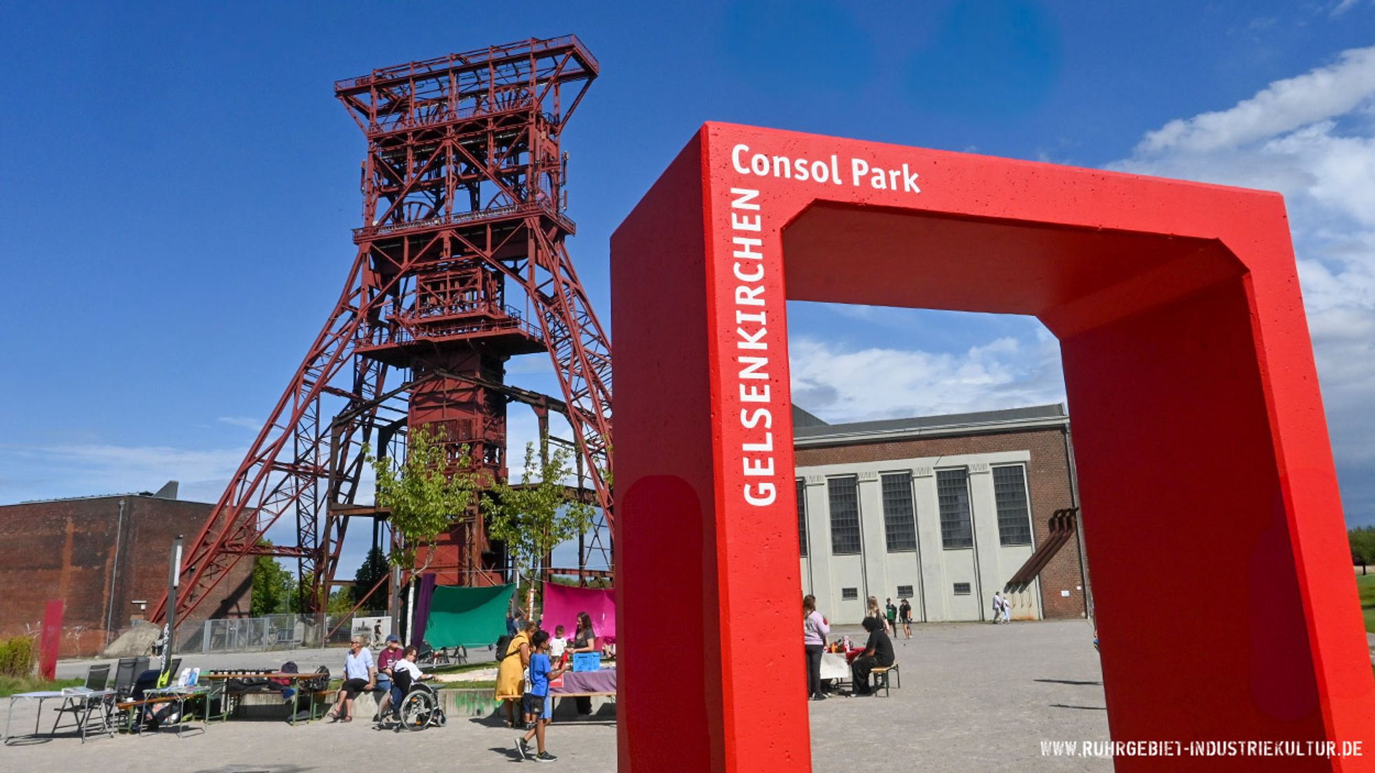 Ein rotes Fördergerüst einer Zeche. Davor ein Platz mit Bäumen. Zugang zum Platz ist ein knallroter Betonrahmen mit der weißen Beschriftung Consol-Park Gelsenkirchen.