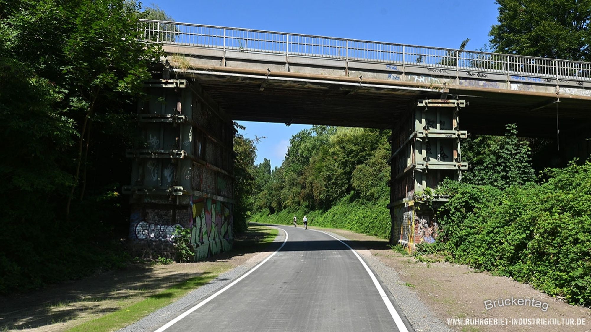 Ein Radweg führt unter einer Brücke hindurch. Die zwei steinernen Pfeiler sind mit einem zusätzlichen Stütz-Gerüst ausgestattet.