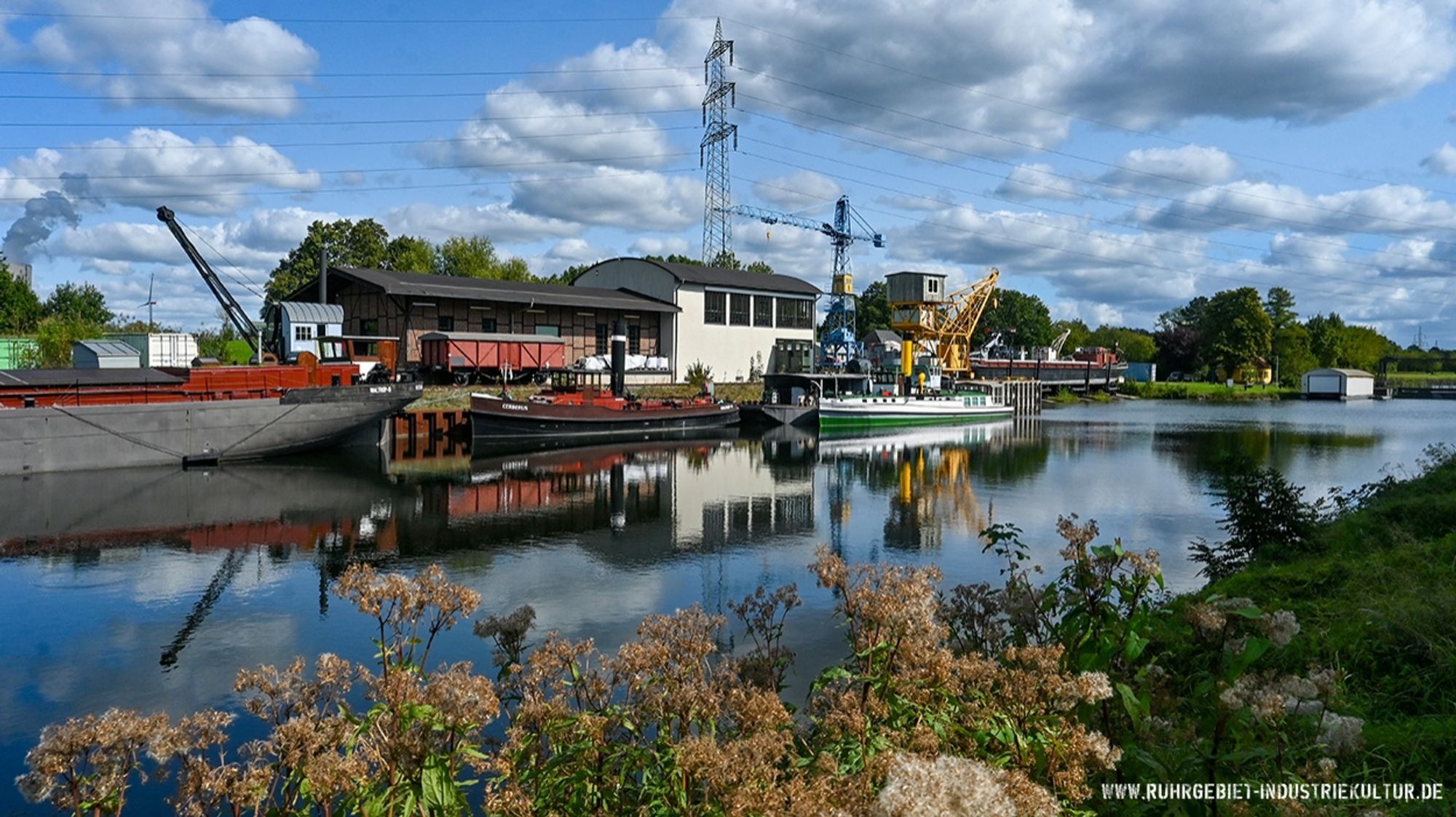 Blick über ein Kanal auf Schiffe auf der anderen Seite, die sich im Wasser spiegeln. Zu sehen sind außerdem Teile einer Hafenanlage mit kleinen Kränen.