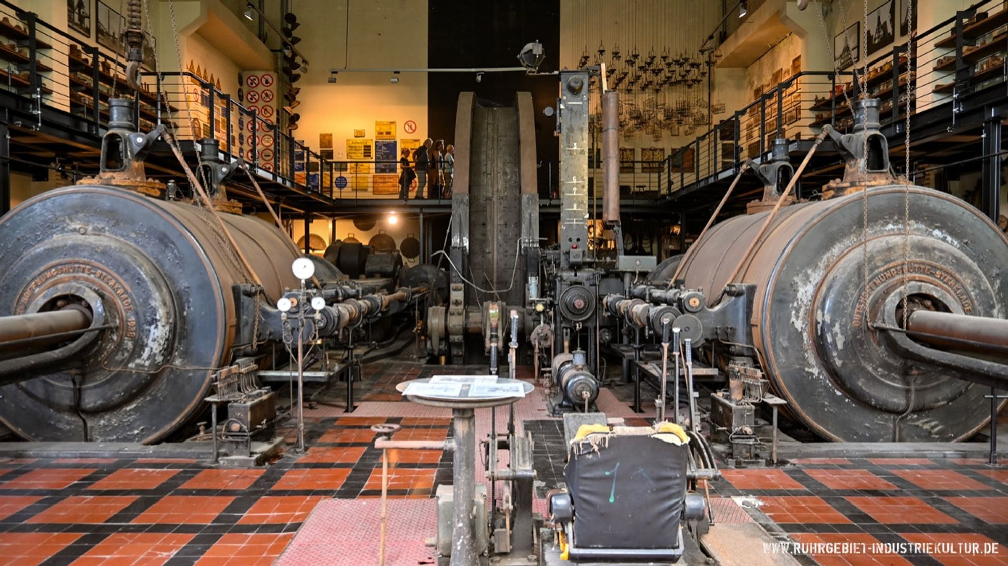 Eine Dampfmaschine in einer Maschinenhalle. Die Halle hat eine Empore, auf der zahlreiche Exponate einer Ausstellung zum Thema Bergbau zu besichtigen sind.