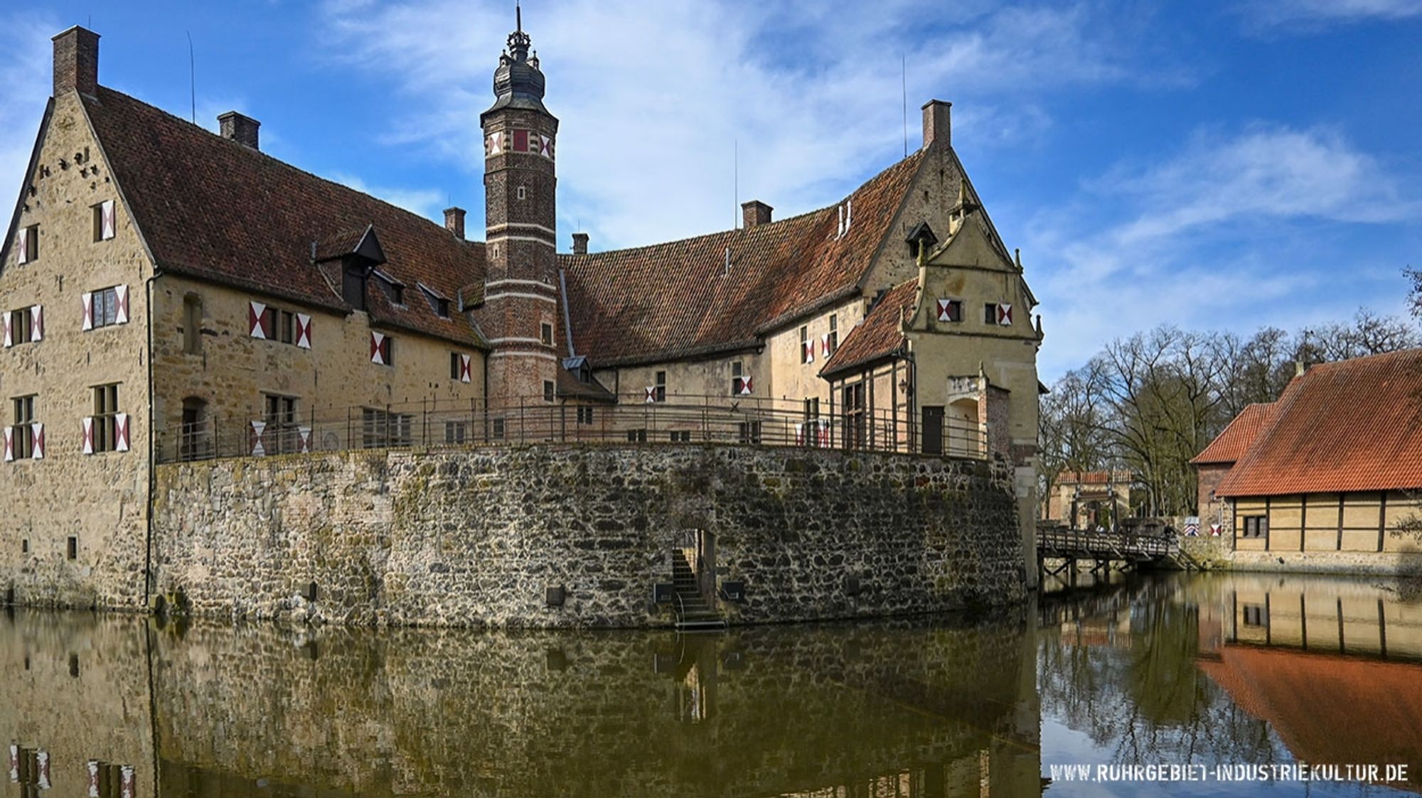 Eine Wasserburg inmitten eines Wassergrabens oder Teiches. Zu sehen ist ein mehreckiger Turm mit Zwiebelhaube, der von zwei Gebäudetrakten mit rot-weißen Fensterläden begrenzt wird. Außerhalb der Burg ist ein Fachwerkhaus Teil der Vorburg.