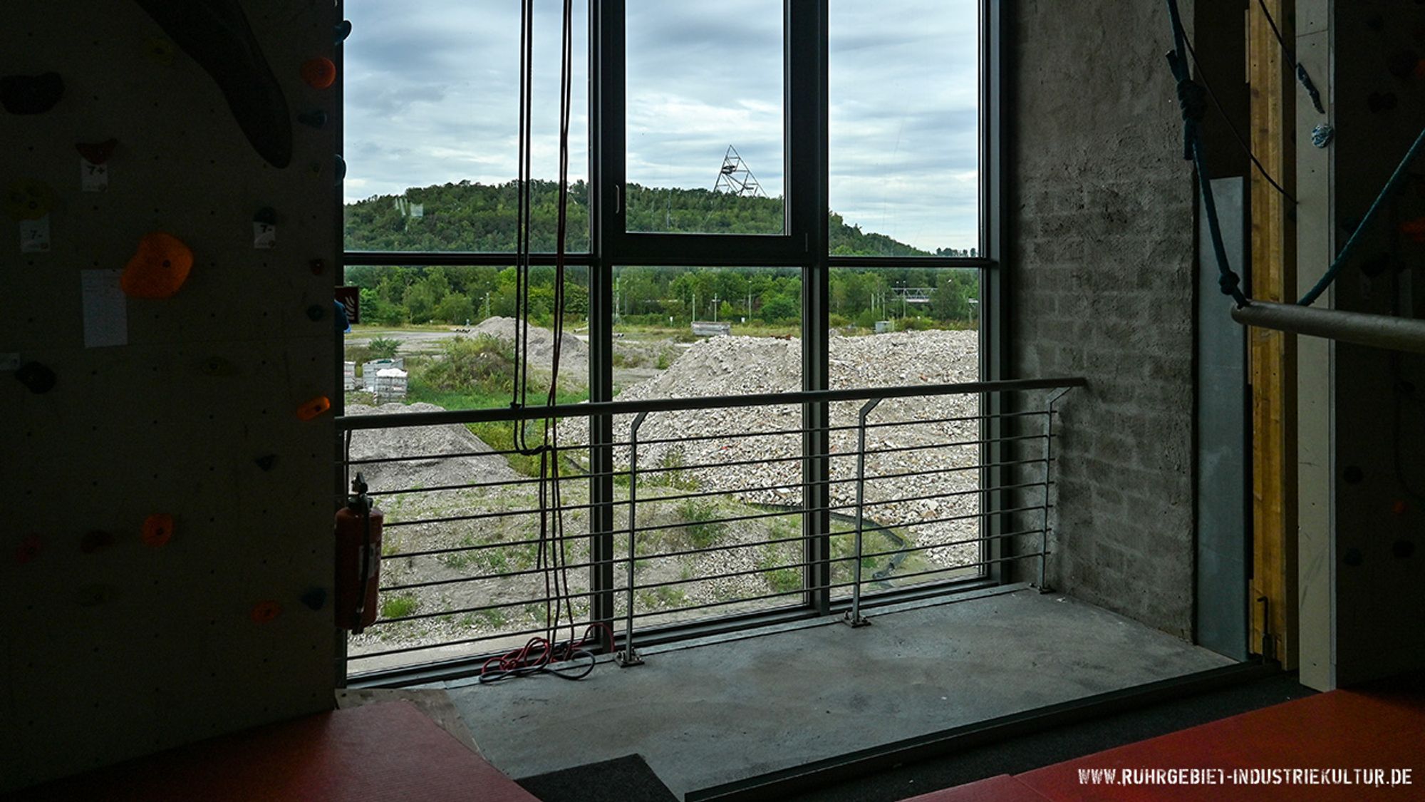 Blick durch ein bodentiefes Fenster auf eine Brachfläche und eine Bergehalde mit dem Tetraeder von Bottrop im Hintergrund.