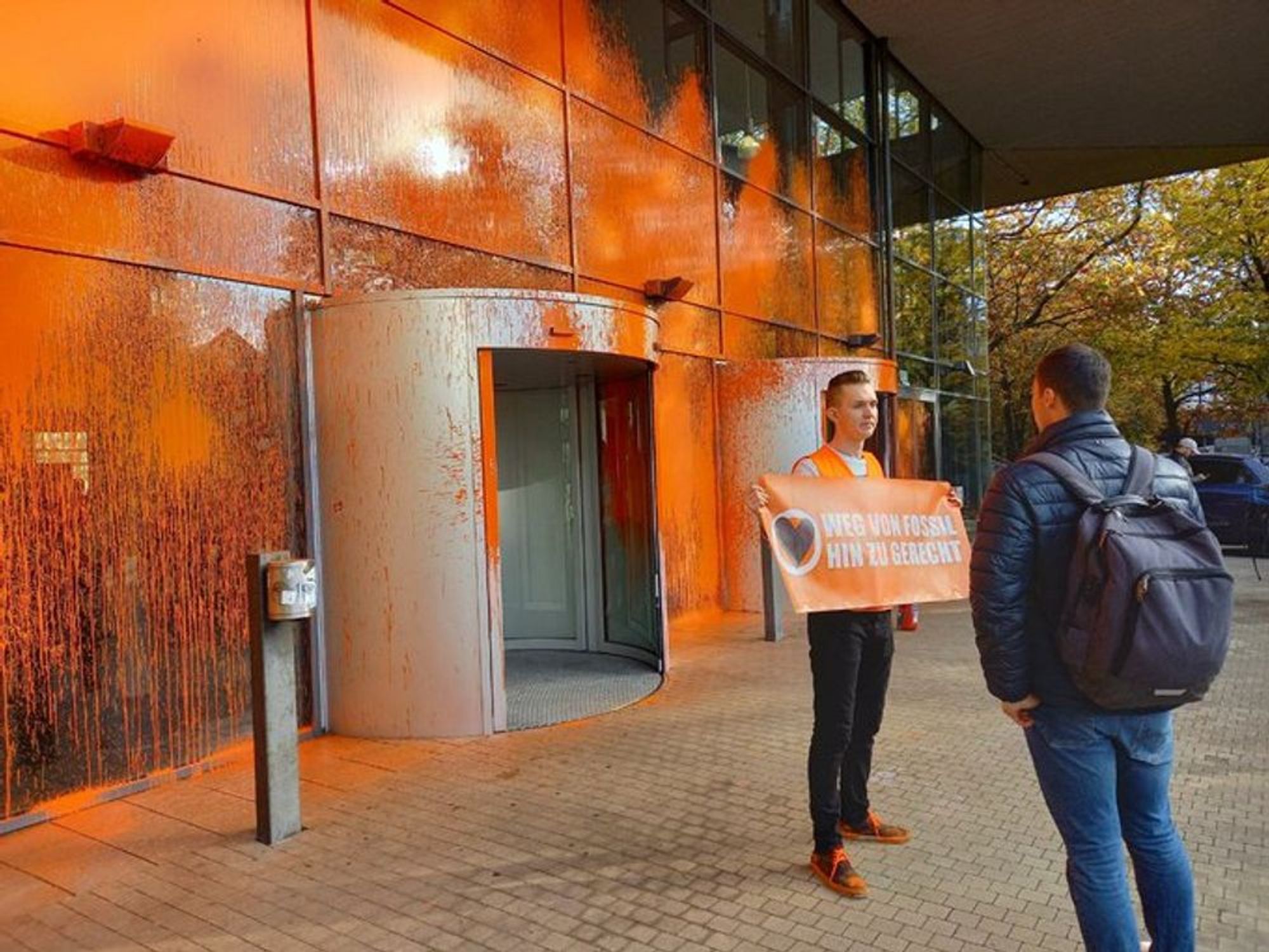 Foto der Letzten Generation. Ein Mensch in Warnweste steht vor einem Gebäude mit Glasfassade, die mit oranger Warnfarbe besprüht wurde, und hält ein Banner auf dem steht: Weg von fossil, hin zu gerecht.