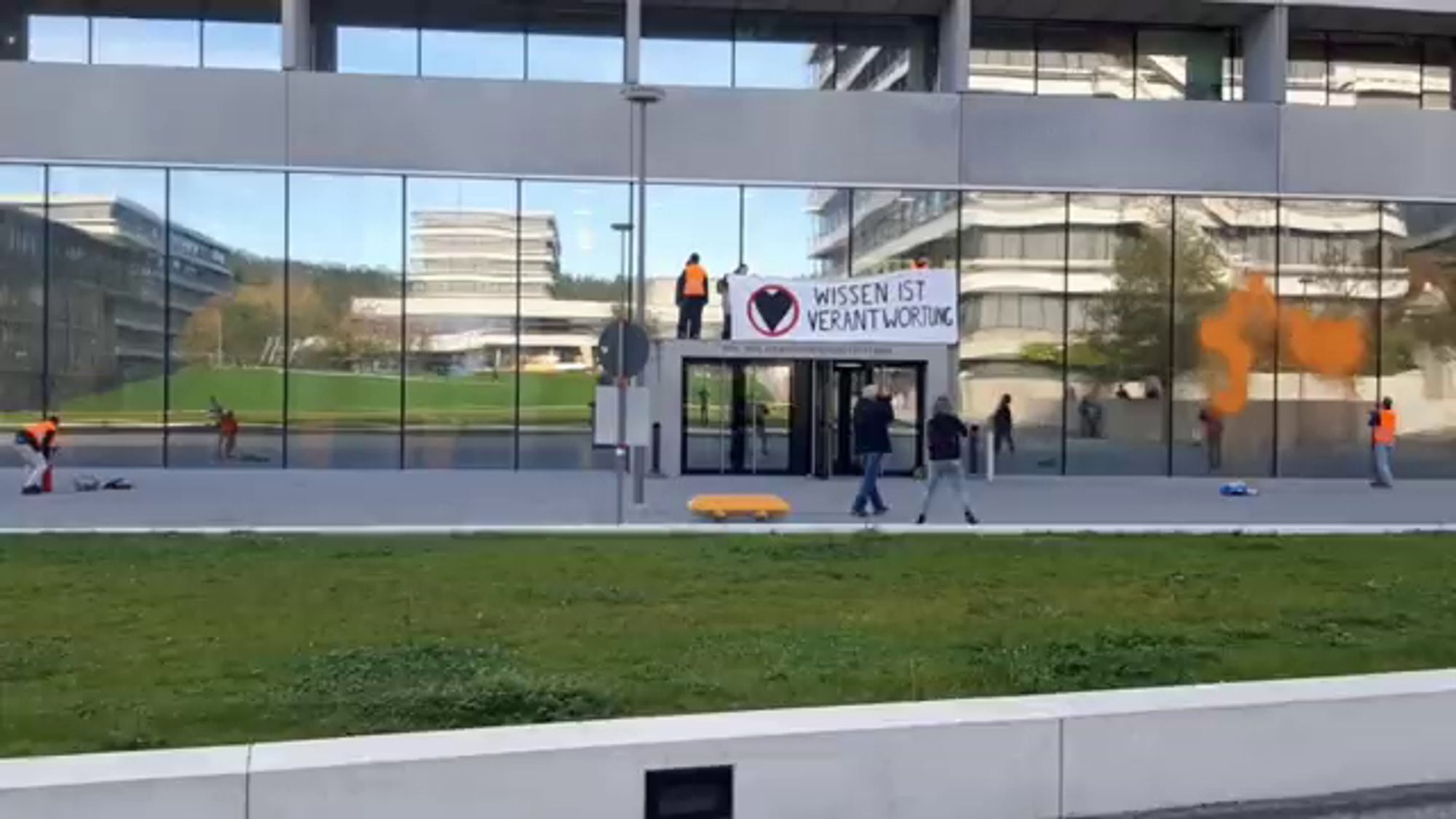 Foto der Letzten Generation. Über dem Eingang der Uni in Tübingen wird ein Banner aufgehängt. Darauf ist das Logo der Letzten Generation und der Text "Wissen ist Verantwortung". Rechts davon sprüht eine Person orangene Farbe an die Fassade.