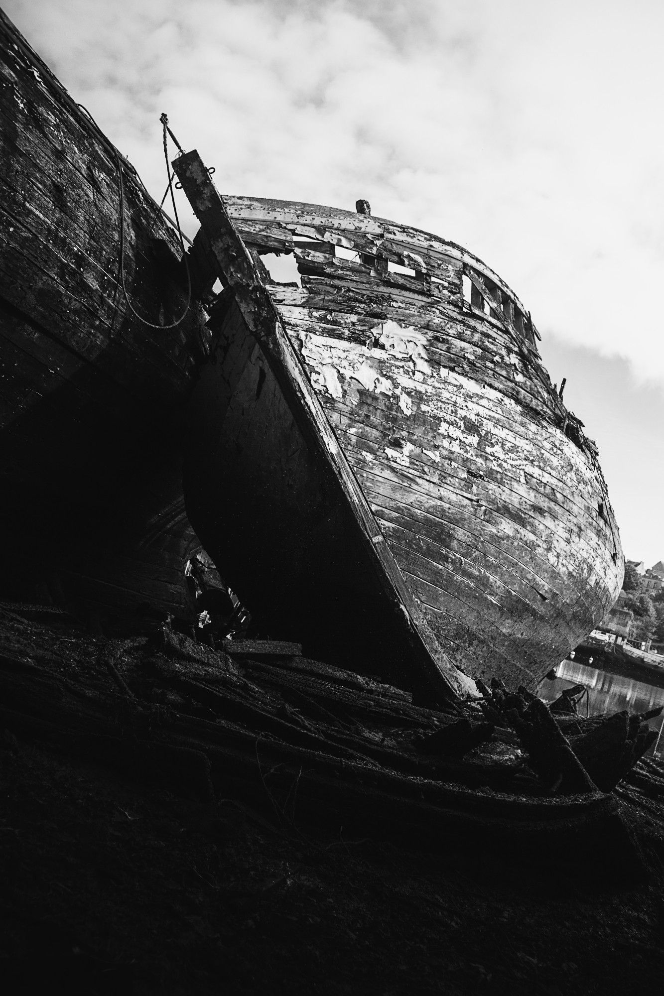 Photographie en noir et blanc de la prou d'un bateau, reposant sur un autre bateau. Le cliché a été pris dans le cimetière de bateaux de Douarnenez. Au premier plan on observe les restes d'une structure de cale. En haut un ciel clair avec des nuages blancs est visible.