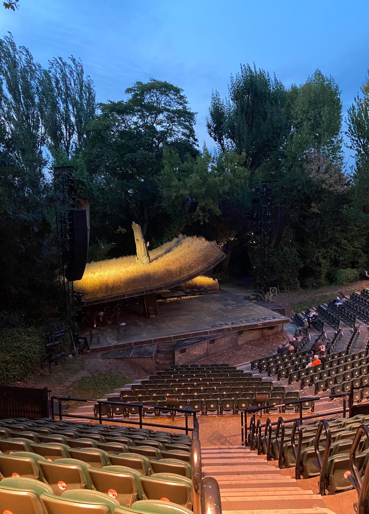 The auditorium at the Open Air Theatre in Regent's Park in London, just after the house opened for this evening's performance (so with the seats mostly empty), with the stage set for their production of Fiddler On The Roof - 

A square wooden stage with the word 'ANATEVKA' (the name of the village where the show takes place) carved into the stage floor. Taking up two-thirds of the stage area, a curved, rising roof with a wheatfield on top and a treetrunk through the middle, mounted high enough that the stage extends underneath it, with room to accommodate the show's orchestra. The stage ringed by trees which form a barrier separating the theatre from the rest of the park.