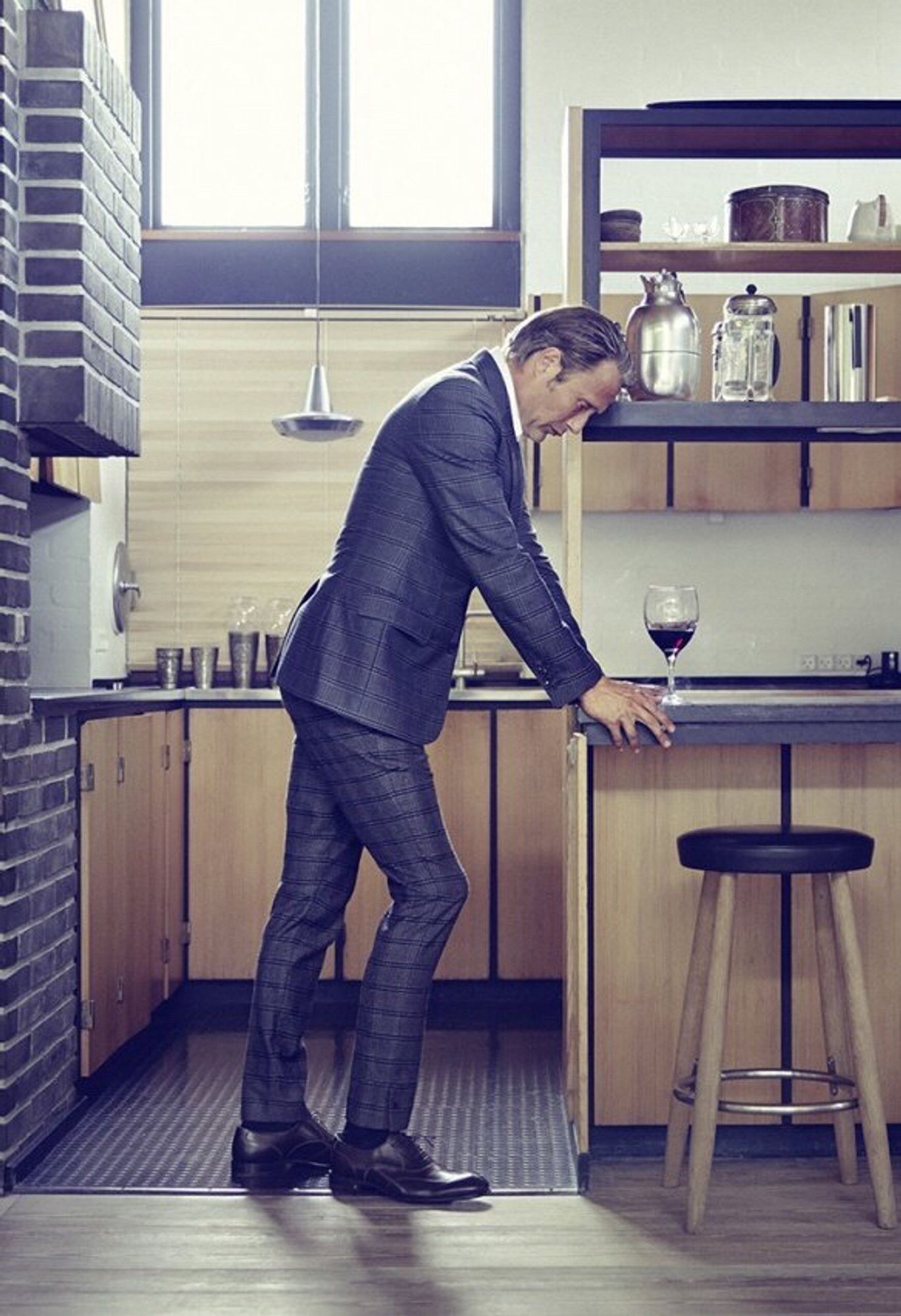 Mads Mikkelsen stands in a smartly decorated kitchen in a dark suit as he braces himself against a kitchen island unit with a glass of red wine in front of him