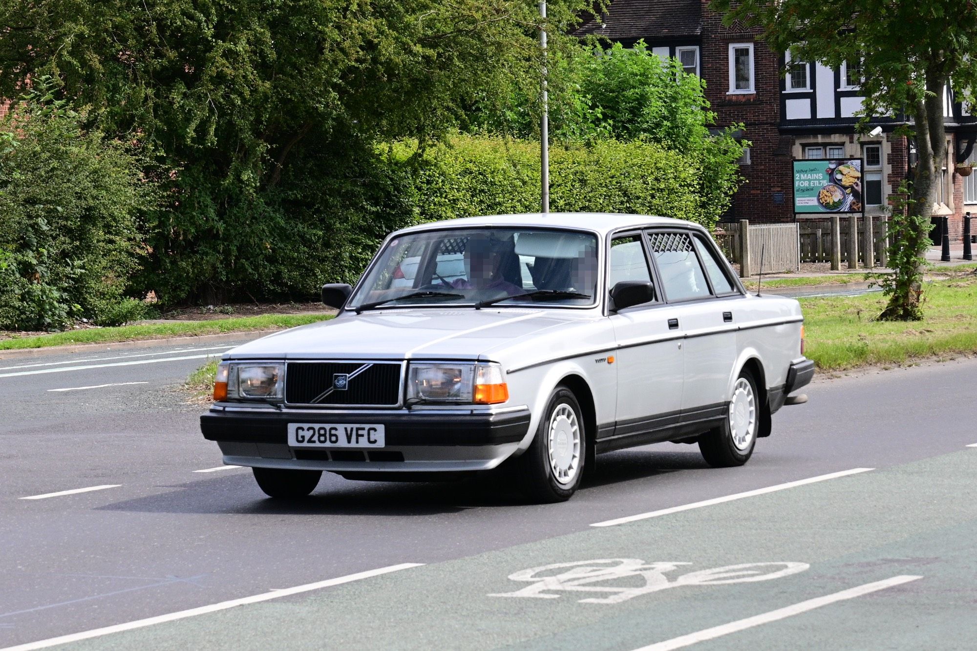 A silver 1990 Volvo 240 four-door sedan on a major road.