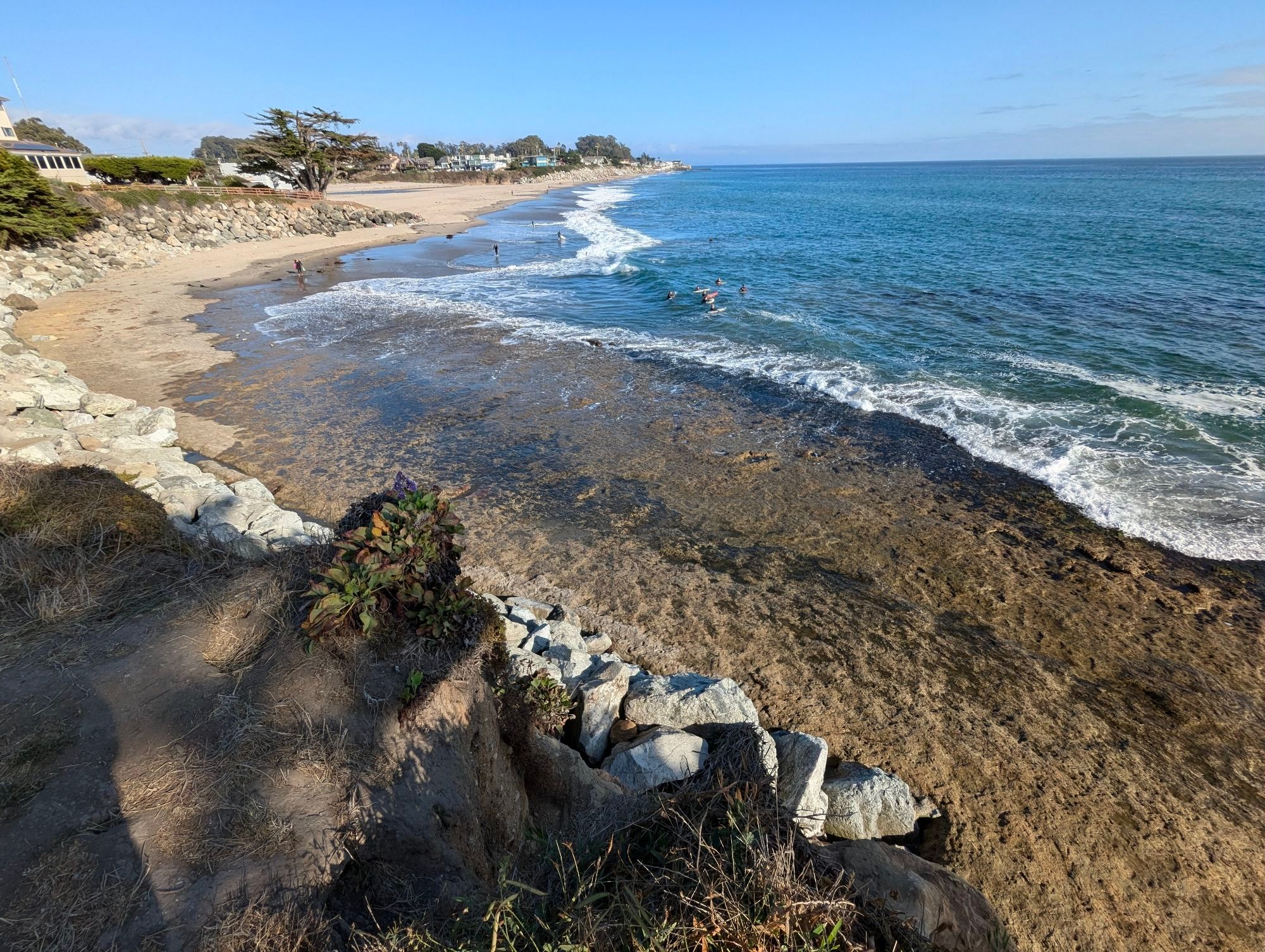 Pacific Coast, beach on the left, blue water on the right, surfers, and a bedrock shelf in the foreground.