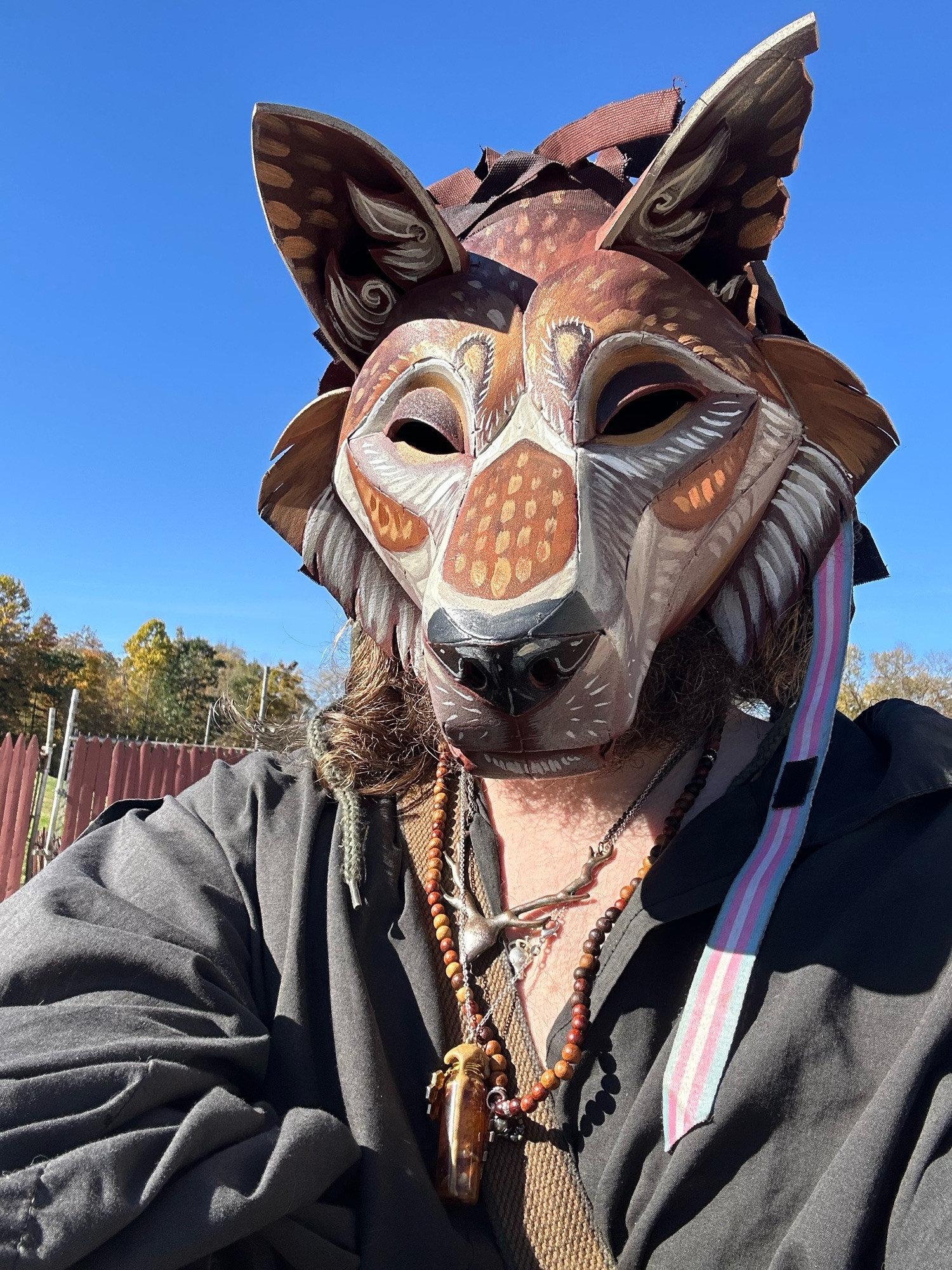 A person in a black shirt with mismatched thrift store jewelry wearing a home-made painted canine mask. 