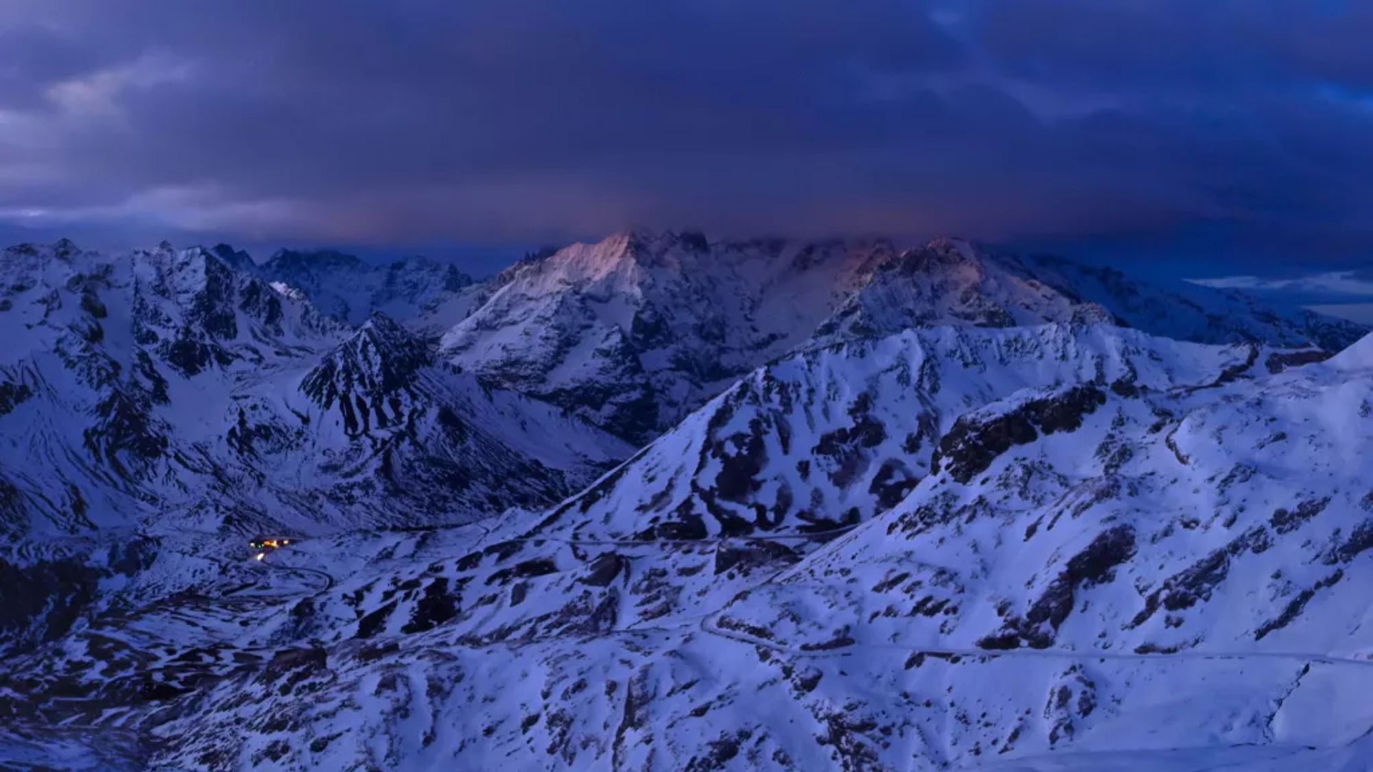 Montagnes enneigées, avec des nuages bas, juste avant le lever du soleil. En tout petit, les éclairages au Col du Lautaret.