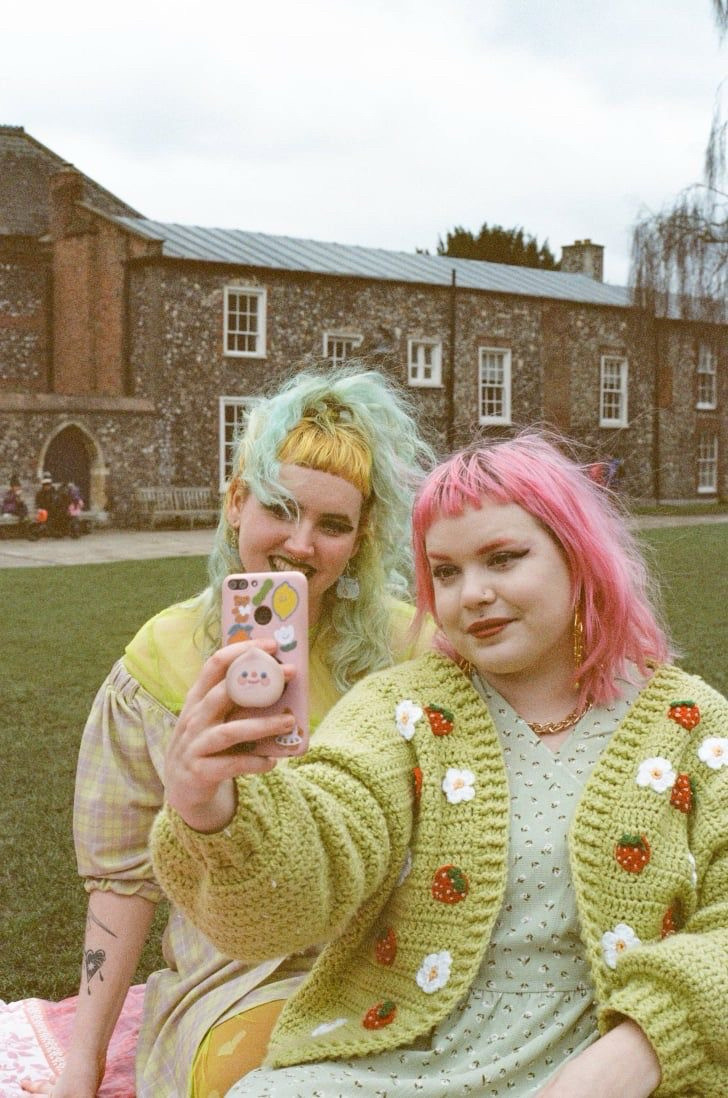 Duas pessoas sentadas em um gramado tirando uma selfie. A pessoa à esquerda tem cabelo verde-claro com franja amarela, enquanto a pessoa à direita tem cabelo rosa e usa um cardigã verde de crochê com detalhes de morangos e margaridas. Ao fundo, há um prédio de tijolos e janelas brancas. Ambas estão sorrindo em um ambiente ao ar livre e descontraído